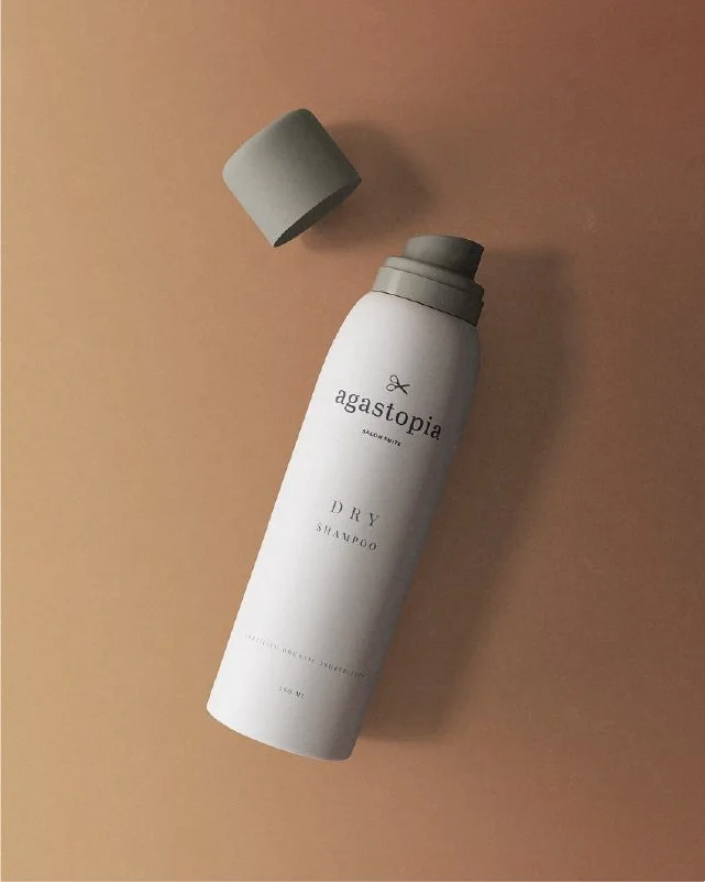 A white bottle of Agastopia Dry Shampoo with a gray cap, placed on a two-tone beige background.