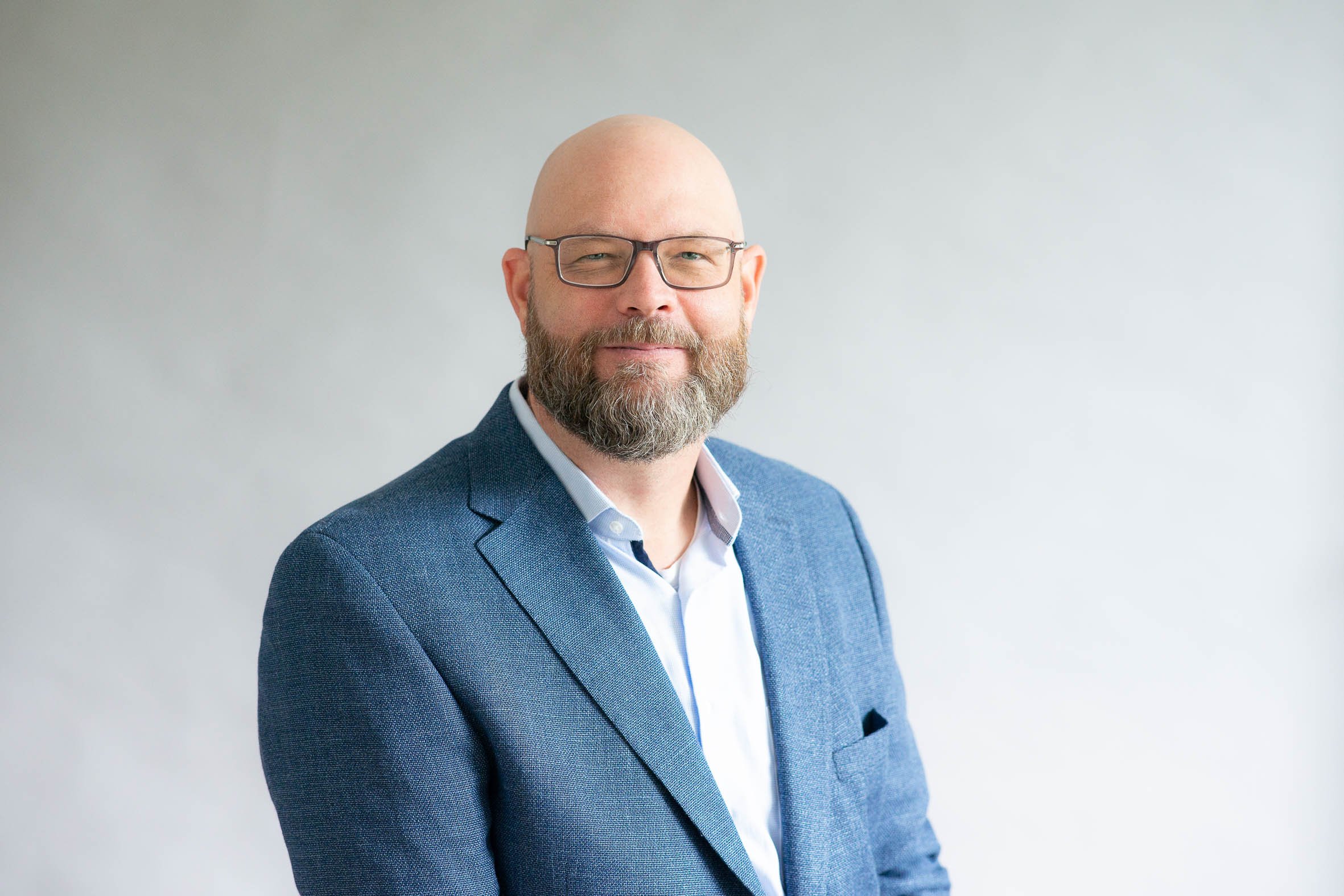 Ein Mann mit Brille, Bart und Kahlkopf, trägt einen blauen Anzug und posiert vor hellem Hintergrund.