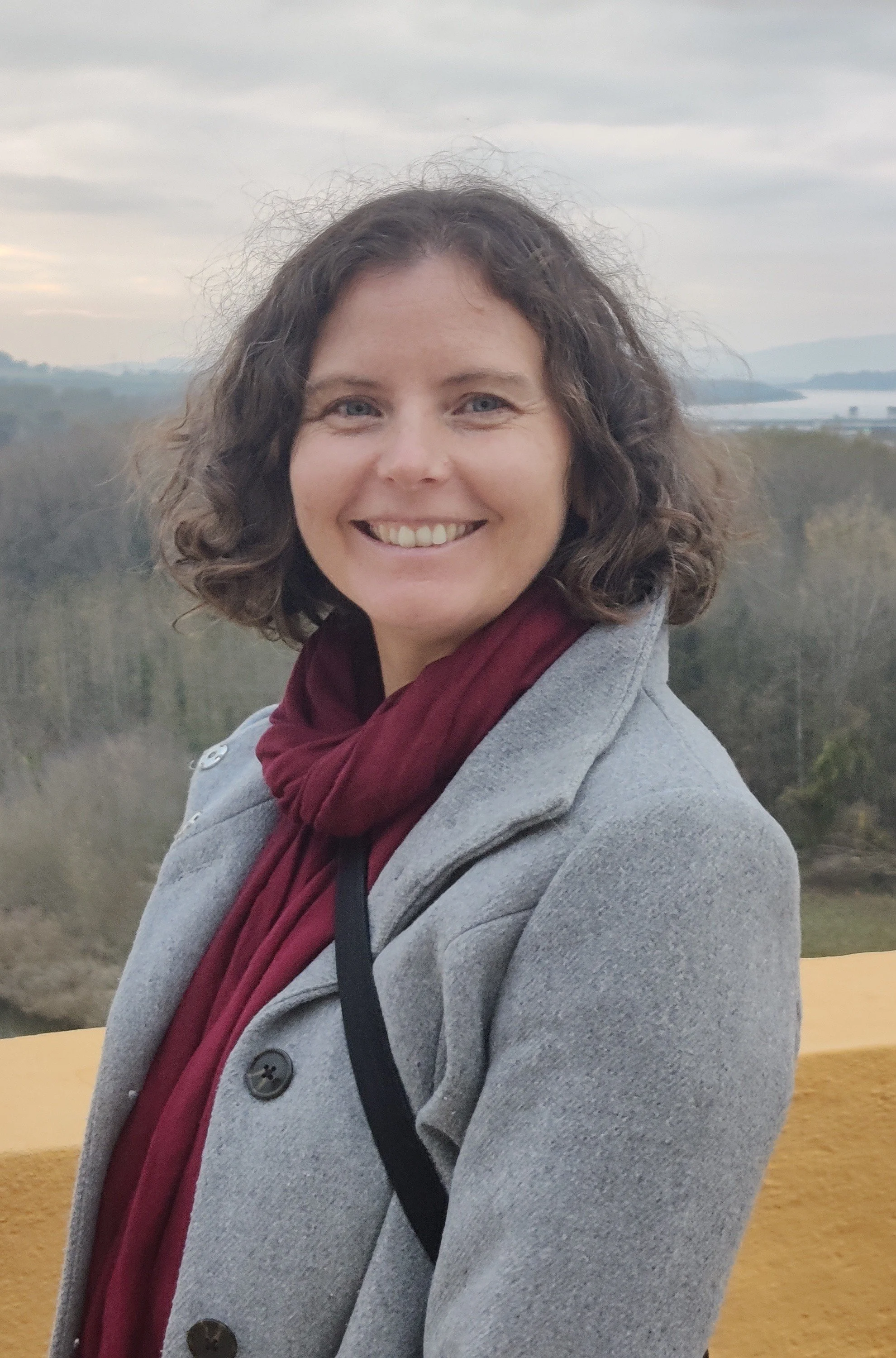 Eine Frau mit kurzen, lockigen braunen Haaren, die lächelnd nach vorne schaut, trägt eine graue Jacke und einen roten Schal, im Hintergrund eine Landschaft mit Bäumen und einem Fluss oder See unter einem bewölkten Himmel.