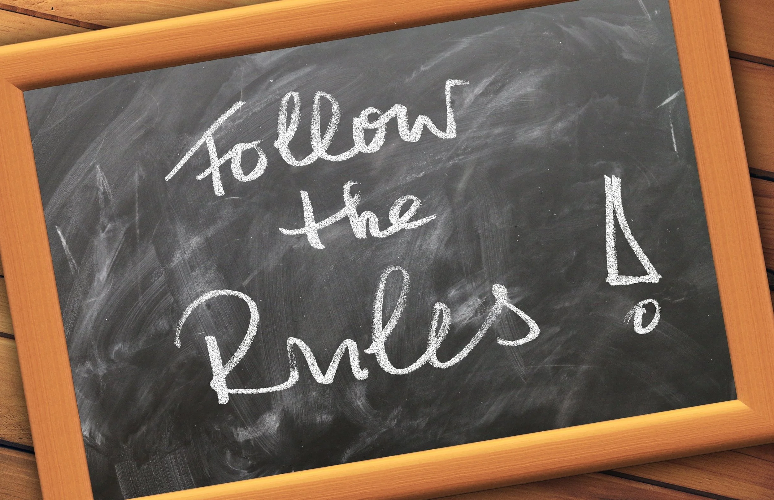 Ein schwarzes Whiteboard mit der handschriftlichen Botschaft 'Follow the Rules' und einem aufgemalten Pfeil nach oben. Das Whiteboard ist von einem Holzrahmen umgeben.