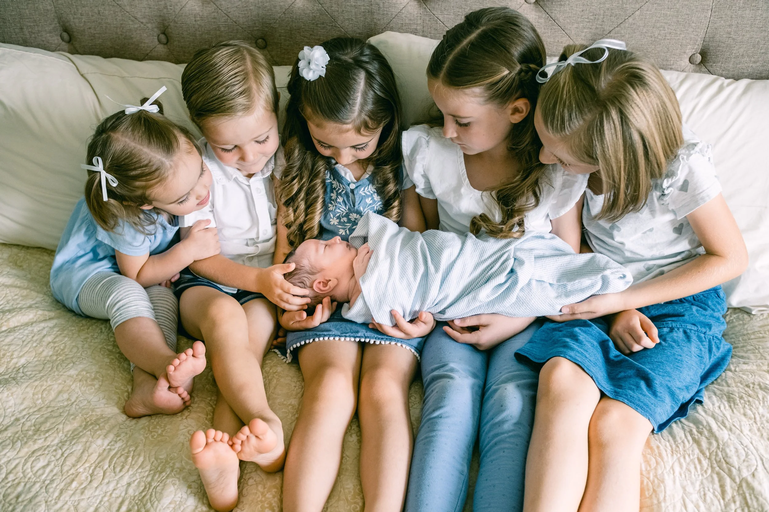 Fünf Mädchen sitzen auf einem Bett und betrachten einen schlafenden kleinen Jungen, der in einer Decke eingewickelt ist, umgeben von Kissen, in einem gemütlichen Schlafzimmer.