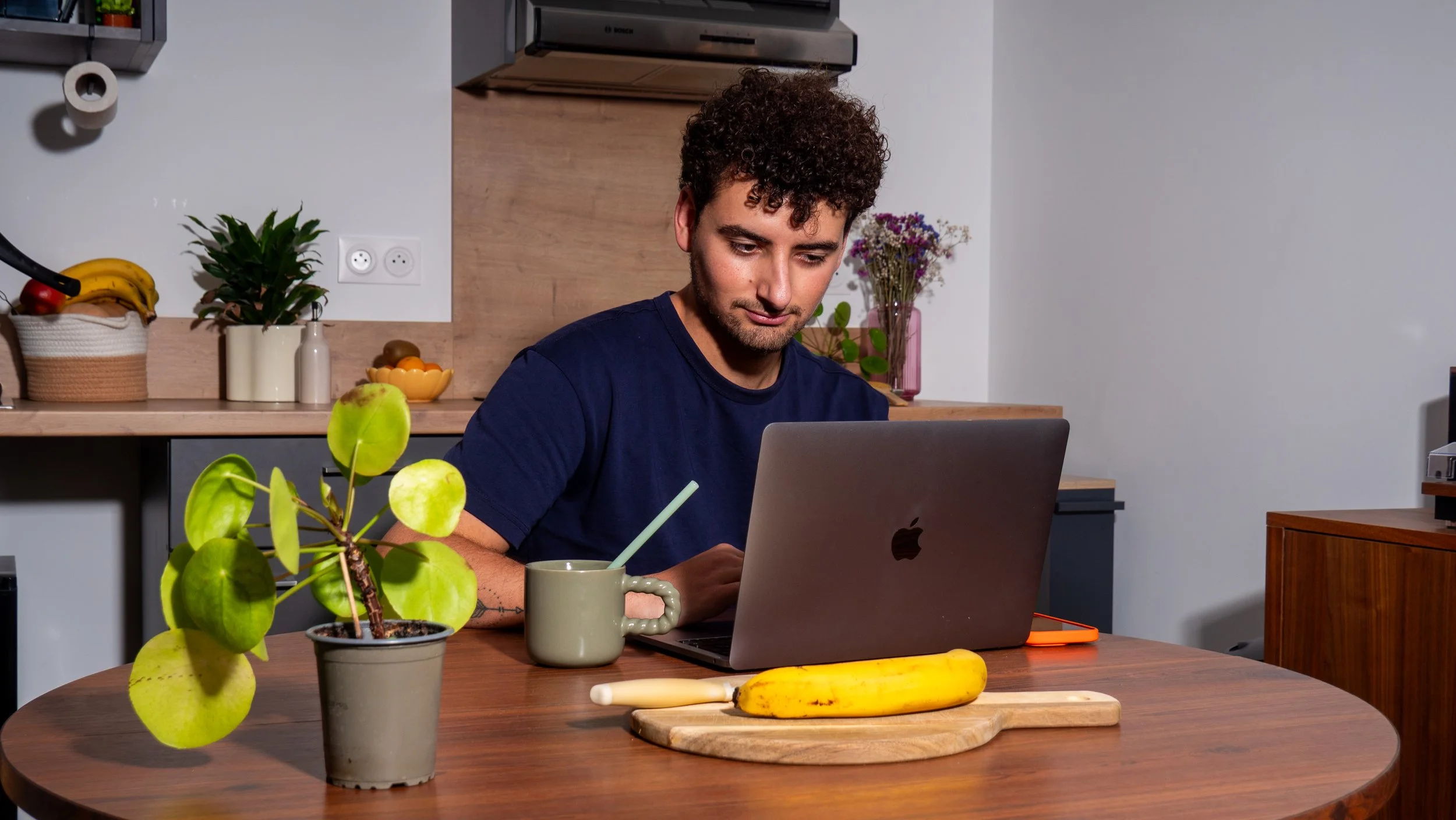 Jeune homme travaillant sur un ordinateur portable à une table en bois, avec une banane, un poireau, une tasse et une plante verte.