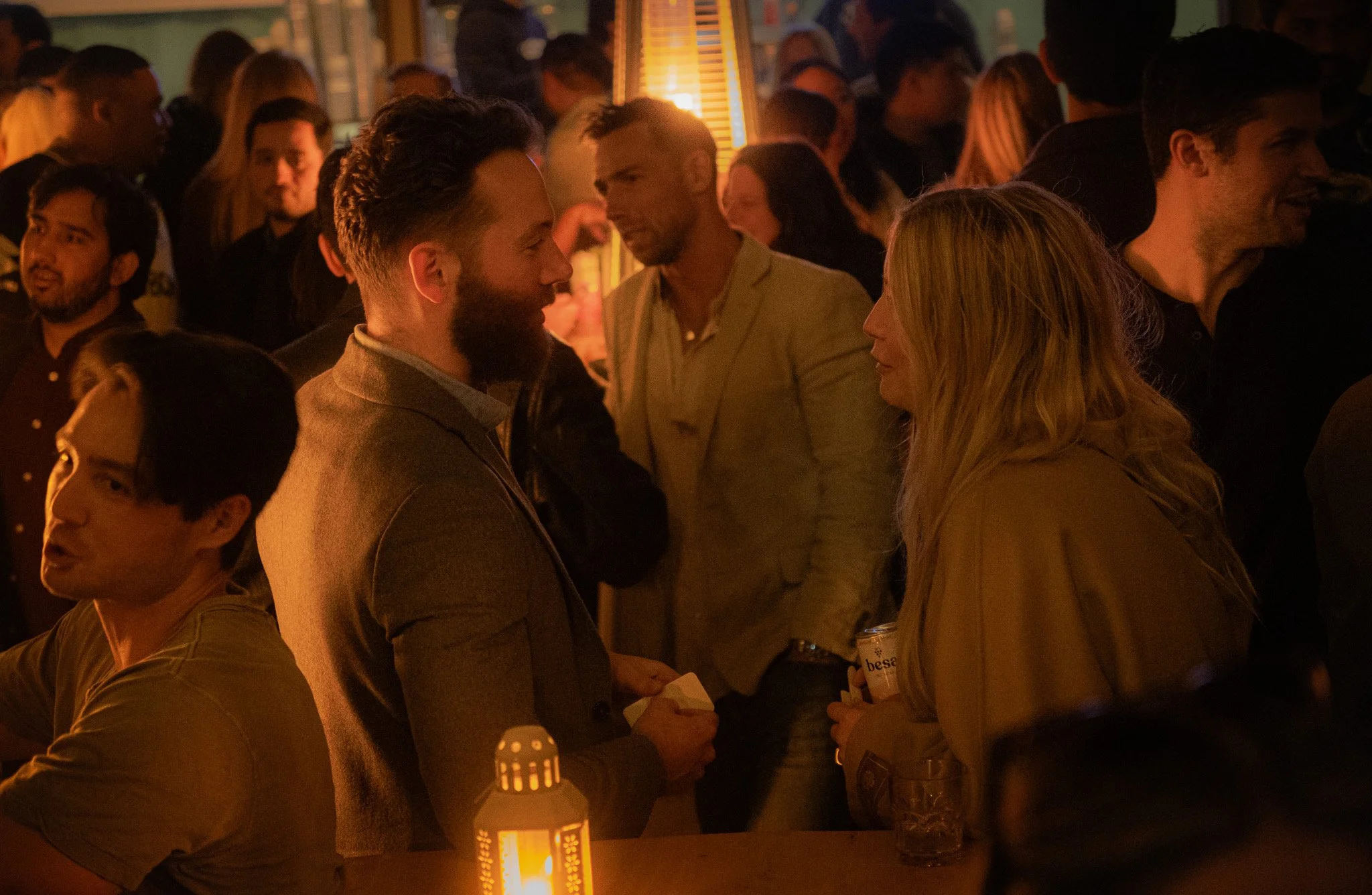 People socializing at a dimly lit indoor event, with a man and woman engaging in conversation, holding drinks, surrounded by other attendees.