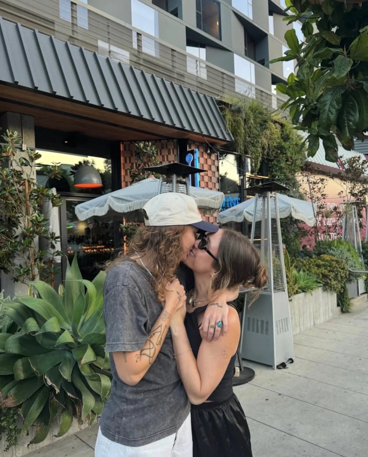 Two women kissing outdoors in front of a restaurant, with plants, patio heaters, and umbrellas in the background.