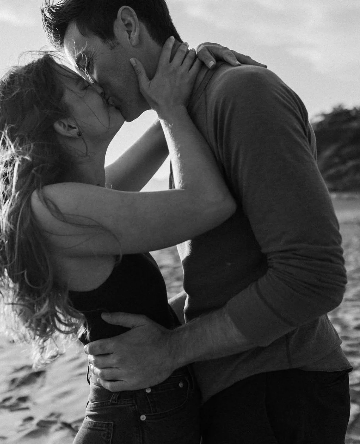 A black-and-white photo of a couple kissing on the beach.