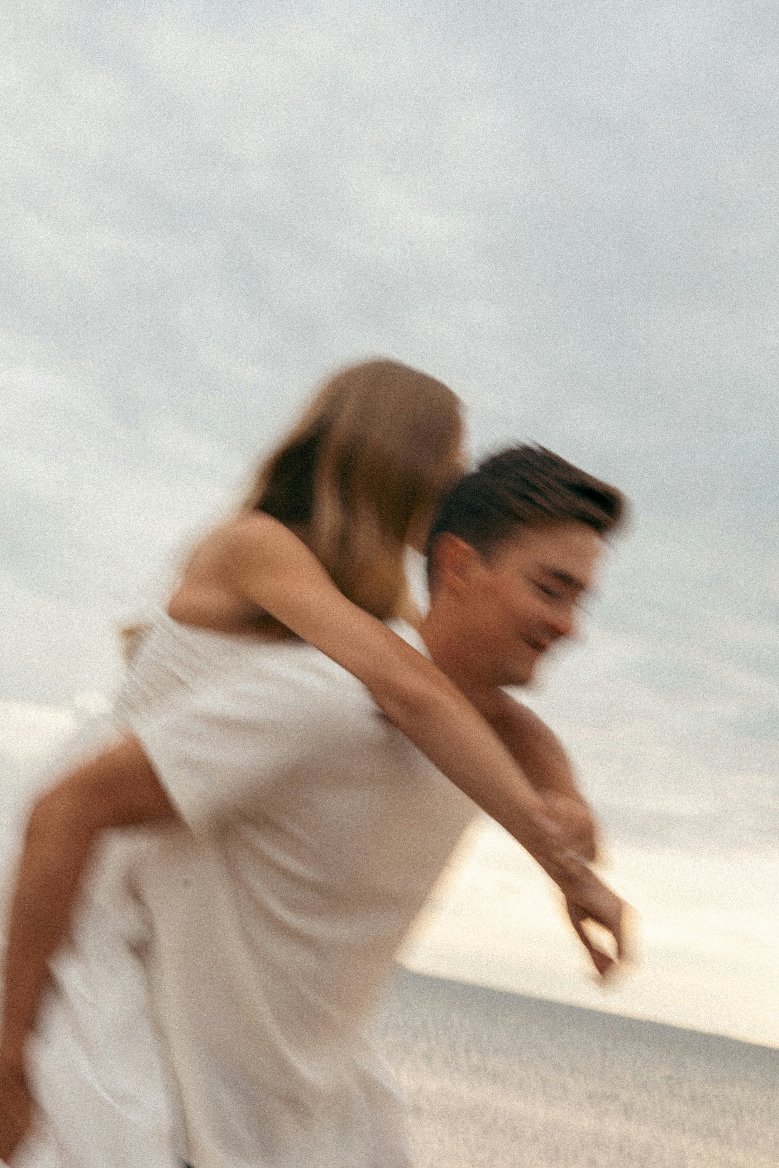 A man giving a piggyback ride to a woman on the beach during sunset, with the sky and ocean in the background.