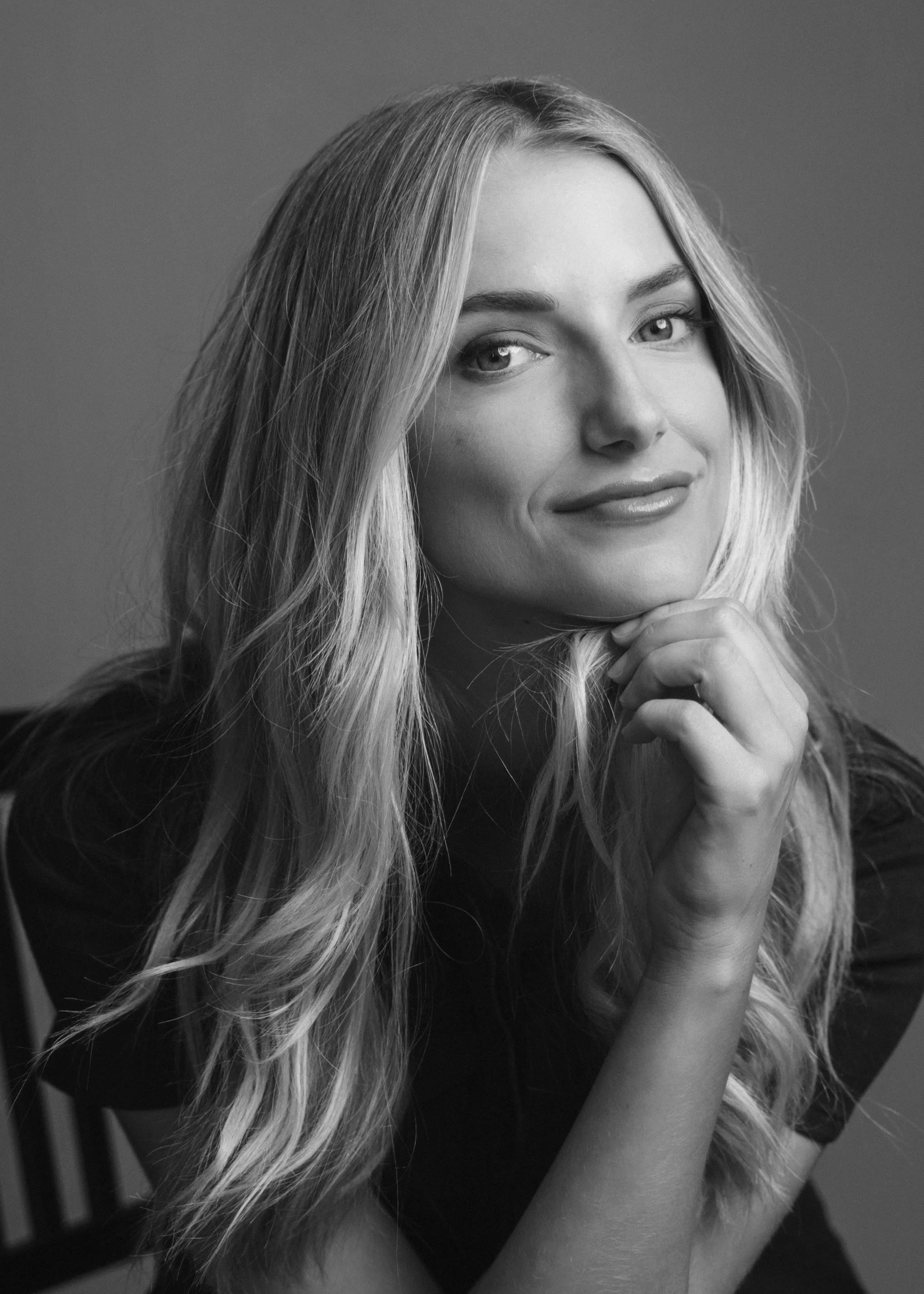 Black and white portrait of a woman with long, wavy hair, smiling and resting her chin on her hand.