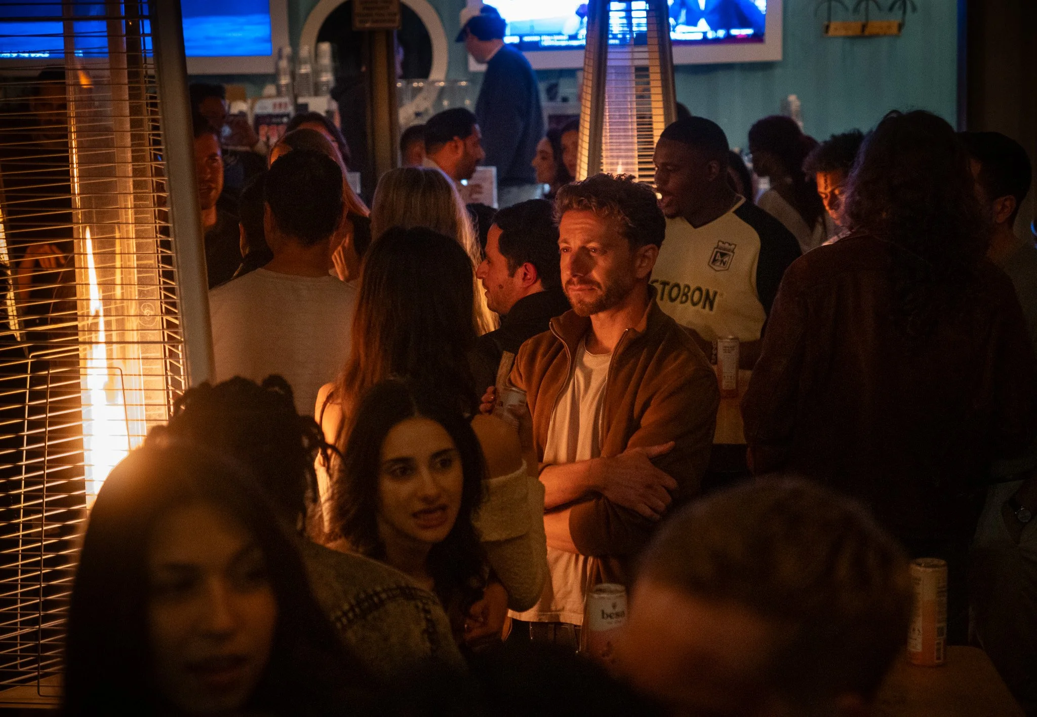 People socializing at a bar or pub with warm lighting, some are talking, and one man is looking pensive, with a television screen in the background.