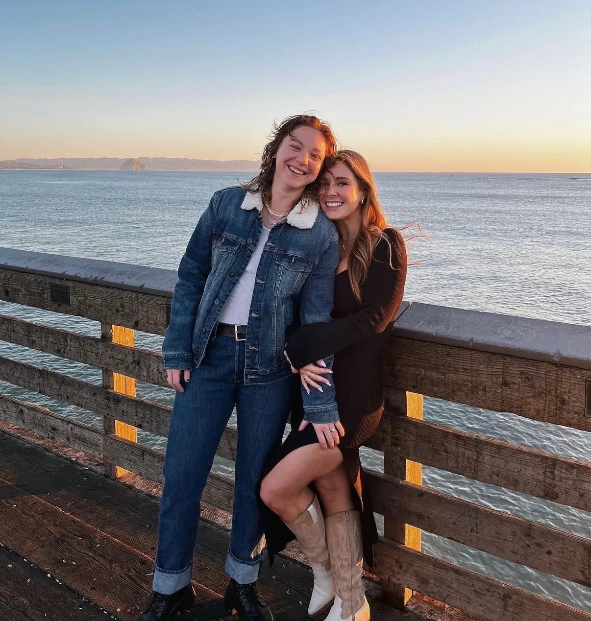 Two women smiling and hugging each other on a pier by the water during sunset, with a distant view of a small island and hills on the horizon.