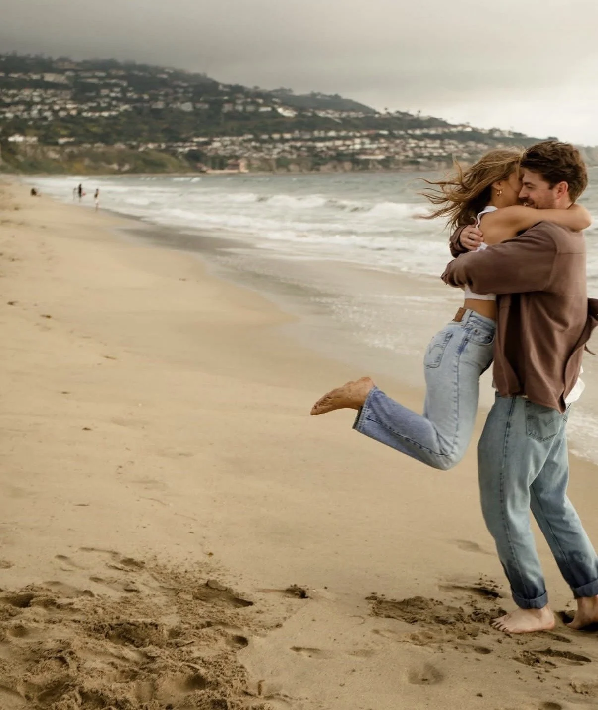 A young couple on a beach, with the man lifting the woman in his arms; she has her arms around his neck, and they are smiling. The beach has sand, ocean waves, and a cloudy sky, with distant hills and houses in the background.