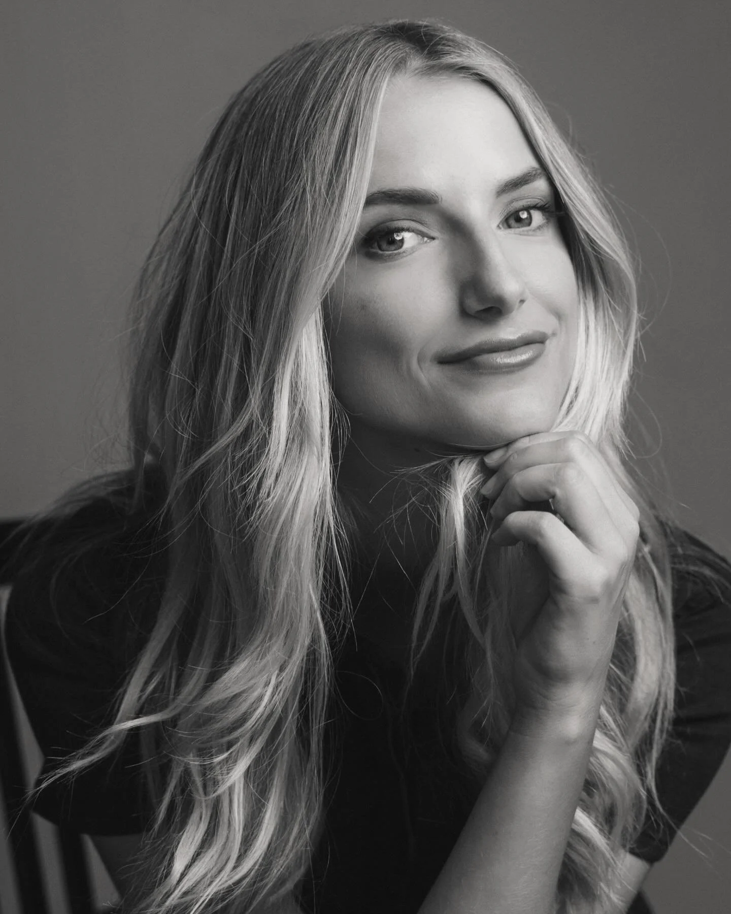 Black and white portrait of a young woman with long wavy hair, smiling gently, with her chin resting on her hand.