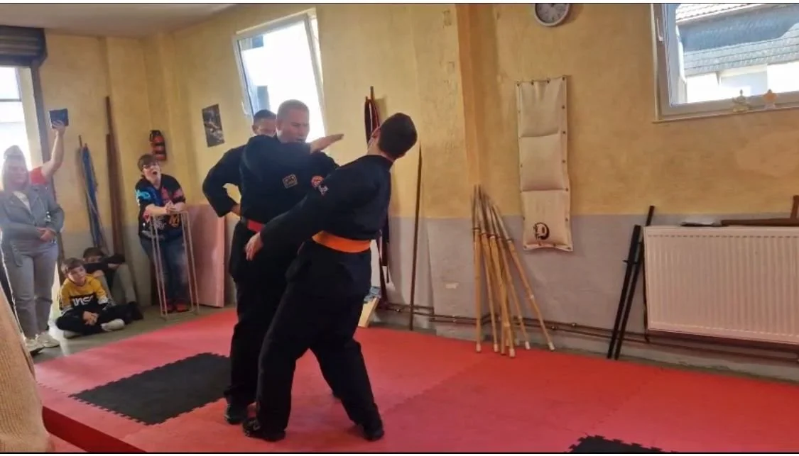 Zwei Martial Arts Kämpfer in schwarzen Gis üben im Trainingsraum, während Zuschauer zuschauen
