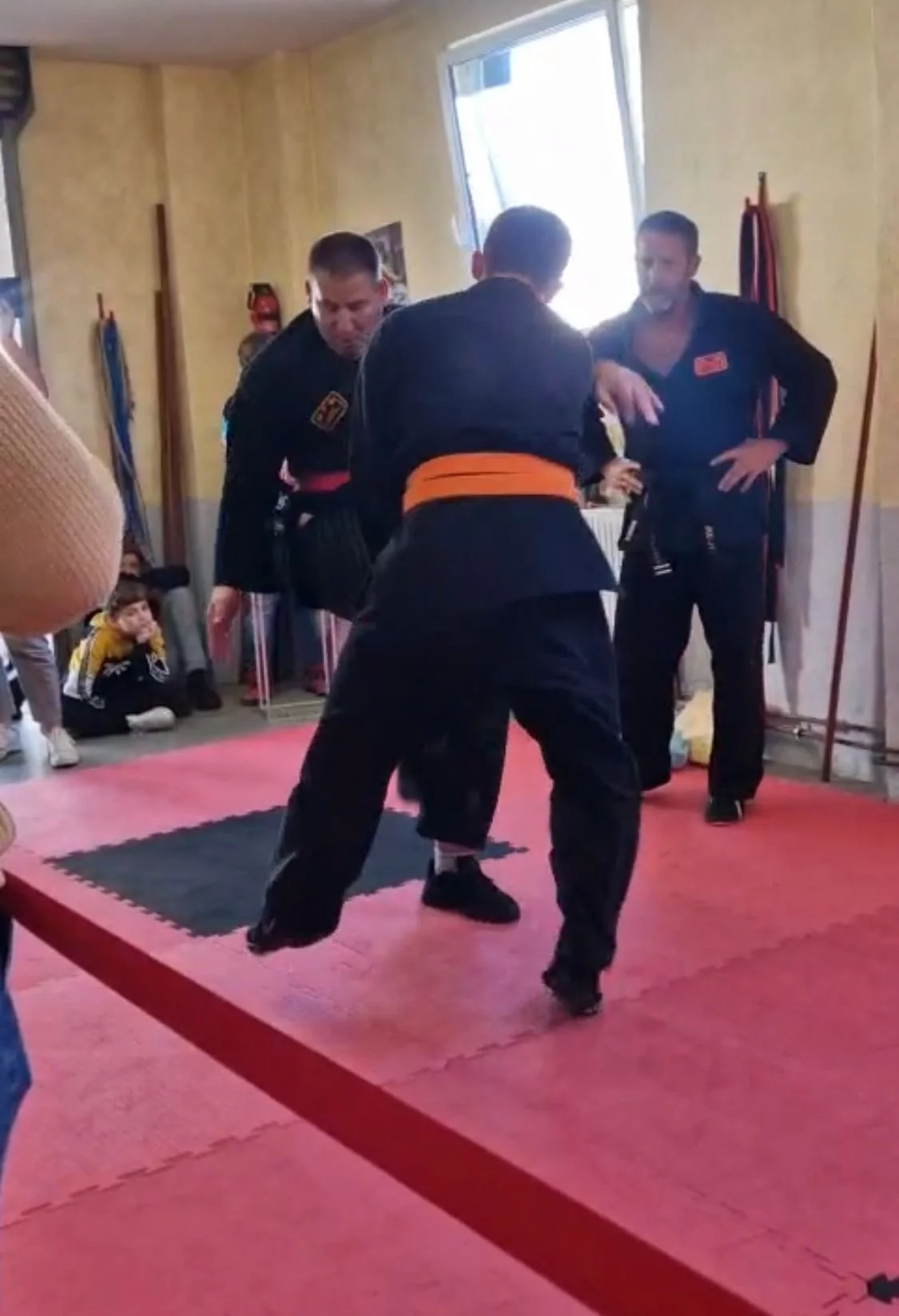 Zwei Kampfsportler im Training auf einem Mattenboden, von einem Trainer beobachtet, in einem Raum mit gelben Wänden und anderen Kindern im Hintergrund.