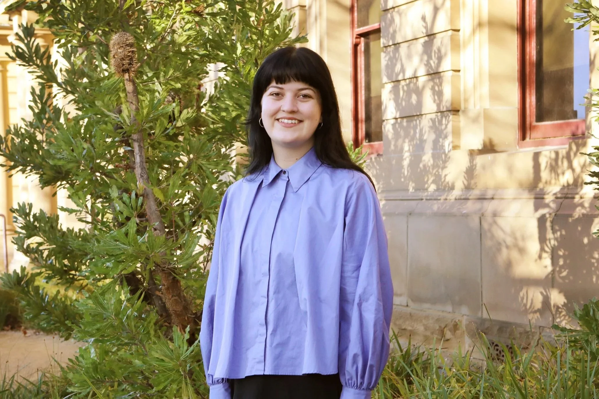 Young woman with dark hair and bangs, smiling, wearing a lavender blouse with puffy sleeves, standing outdoors near green bushes and a yellow building with red window frames.