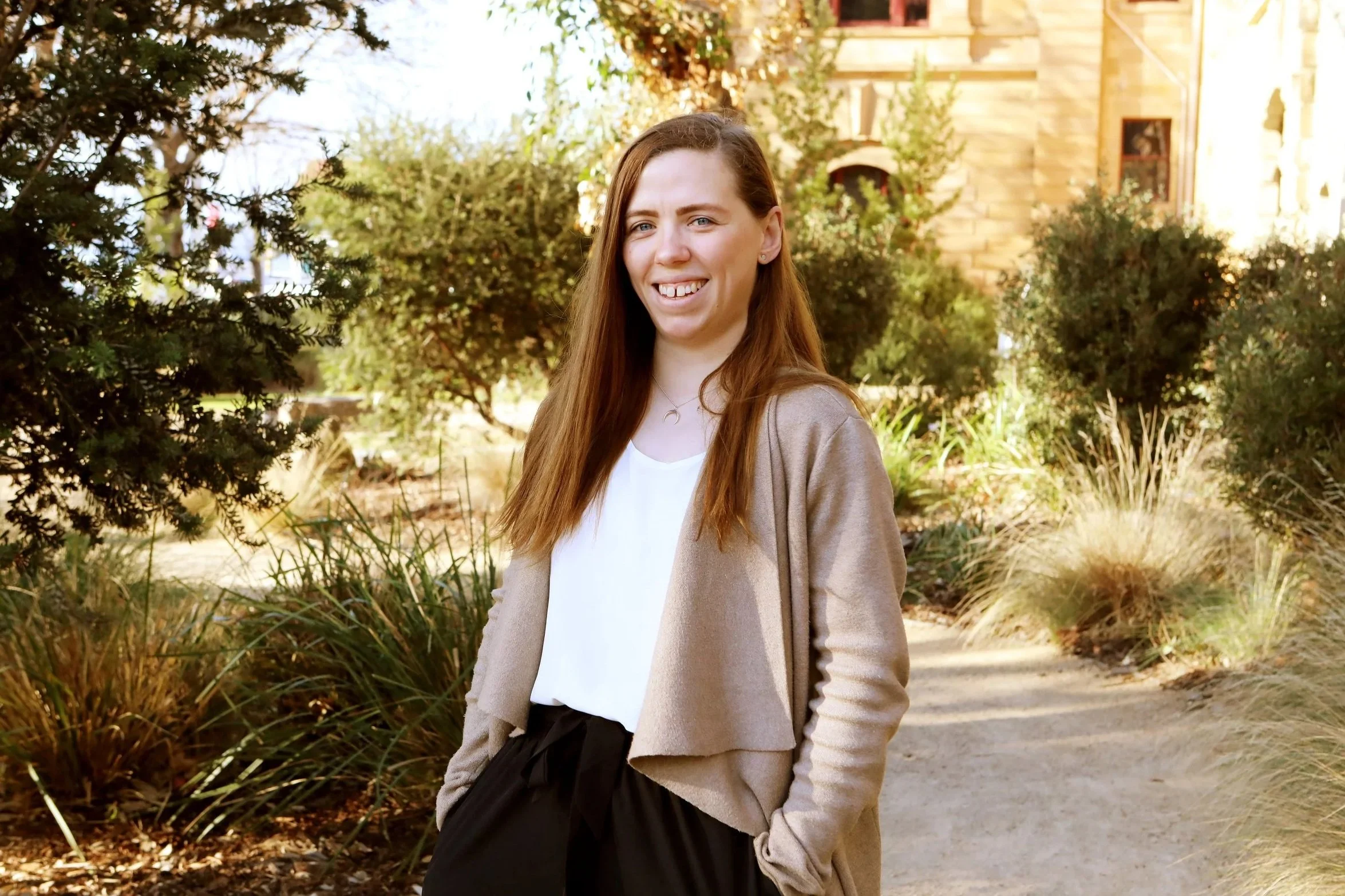 A woman with long reddish hair, smiling, standing outdoors on a garden path with plants and trees in the background.