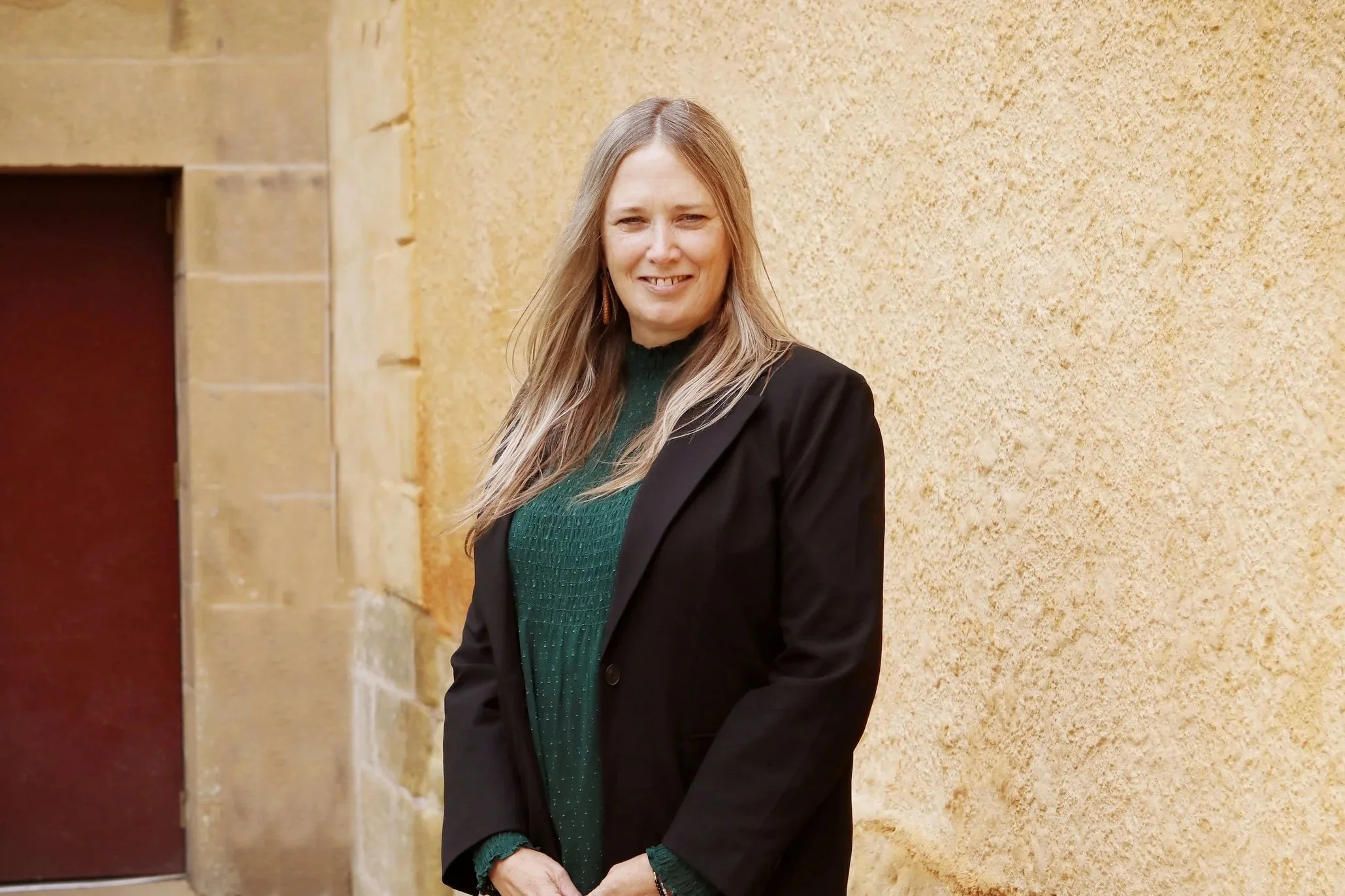 A woman with long blonde hair smiling and standing in front of a yellow brick wall, wearing a black blazer over a green sweater.