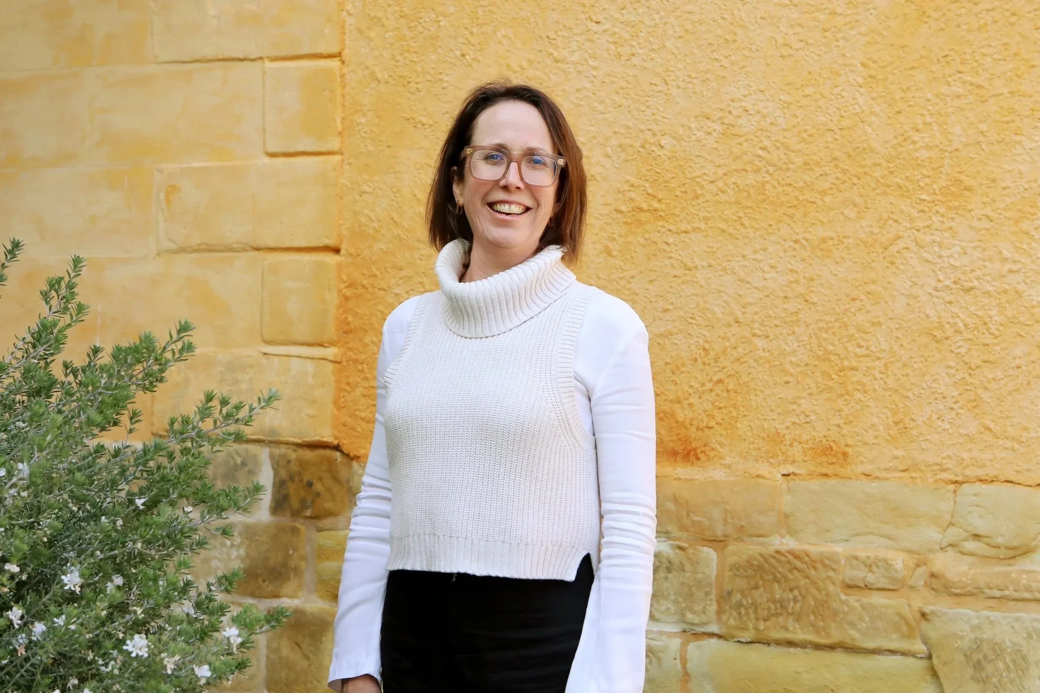 A woman with glasses, shoulder-length brown hair, wearing a white turtleneck sweater vest over a white long sleeve shirt, smiling and standing outdoors near a yellow textured stone wall with greenery in the foreground.