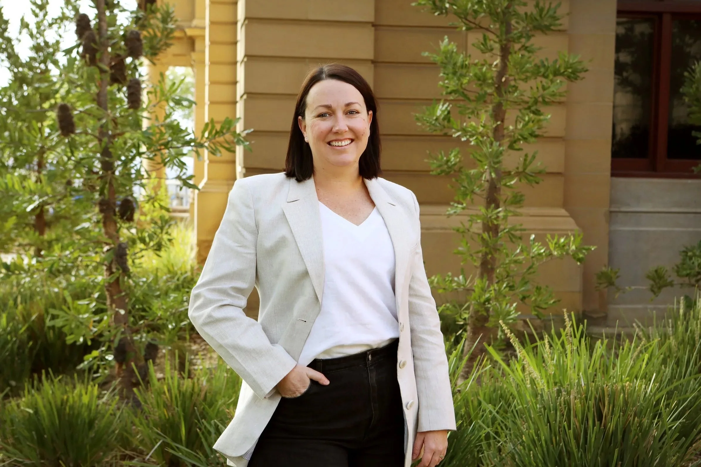 A woman with dark hair smiling outdoors in front of a wooden house, wearing a white blazer, white shirt, and black pants, with green bushes and trees around.