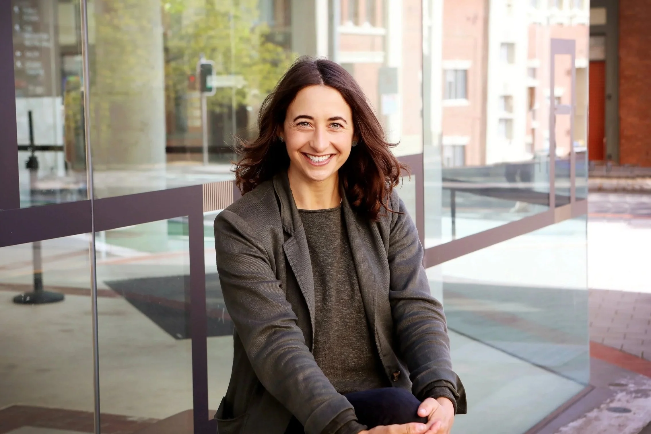 A woman with shoulder-length brown hair sitting outside near a large glass window, smiling at the camera, wearing a dark jacket and shirt, with an urban background of buildings and trees.