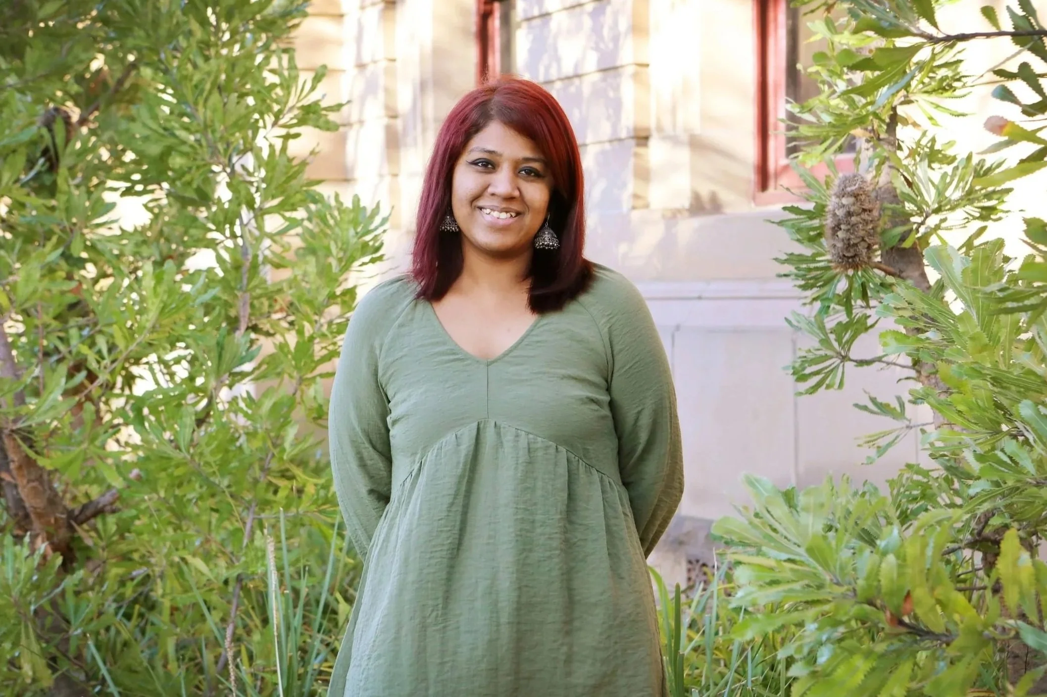 A woman with shoulder-length hair dyed red, wearing a green dress and earrings, standing outdoors amidst green foliage and plants, smiling at the camera.