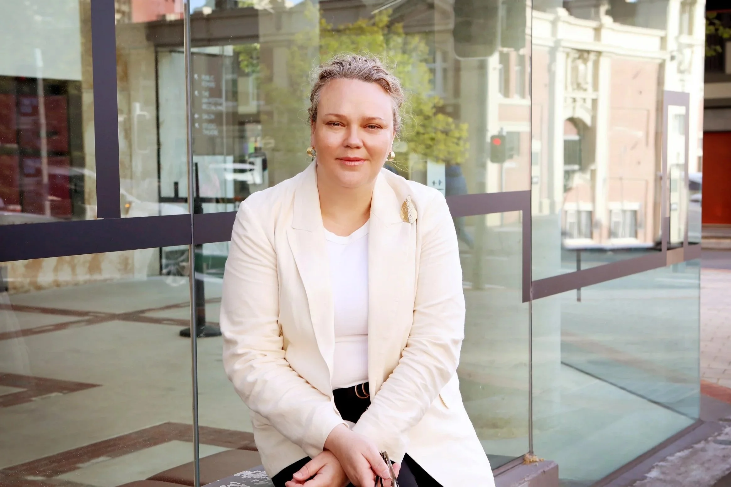 A woman with blonde curly hair, wearing a white blazer with a pin, sits on a bench outside a glass building with reflections of city streets and trees.