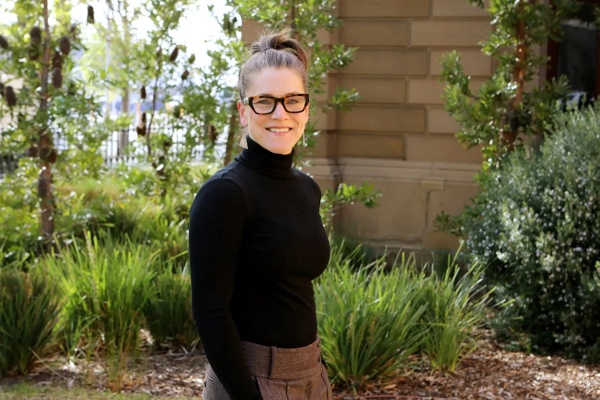 A woman with glasses and a high ponytail, wearing a black turtleneck and gray pants, standing outdoors in front of green bushes and trees, smiling at the camera.