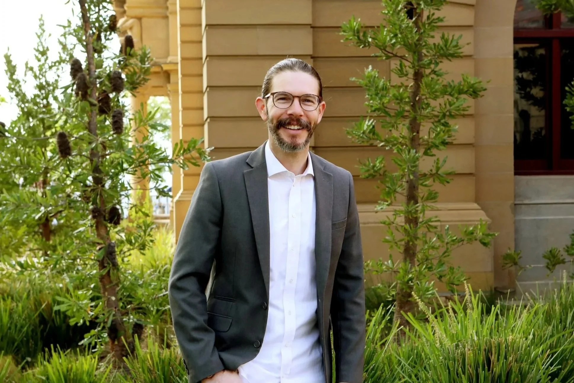 Man with glasses, beard, in a gray suit and white shirt, smiling outdoors near a building with trees and plants.