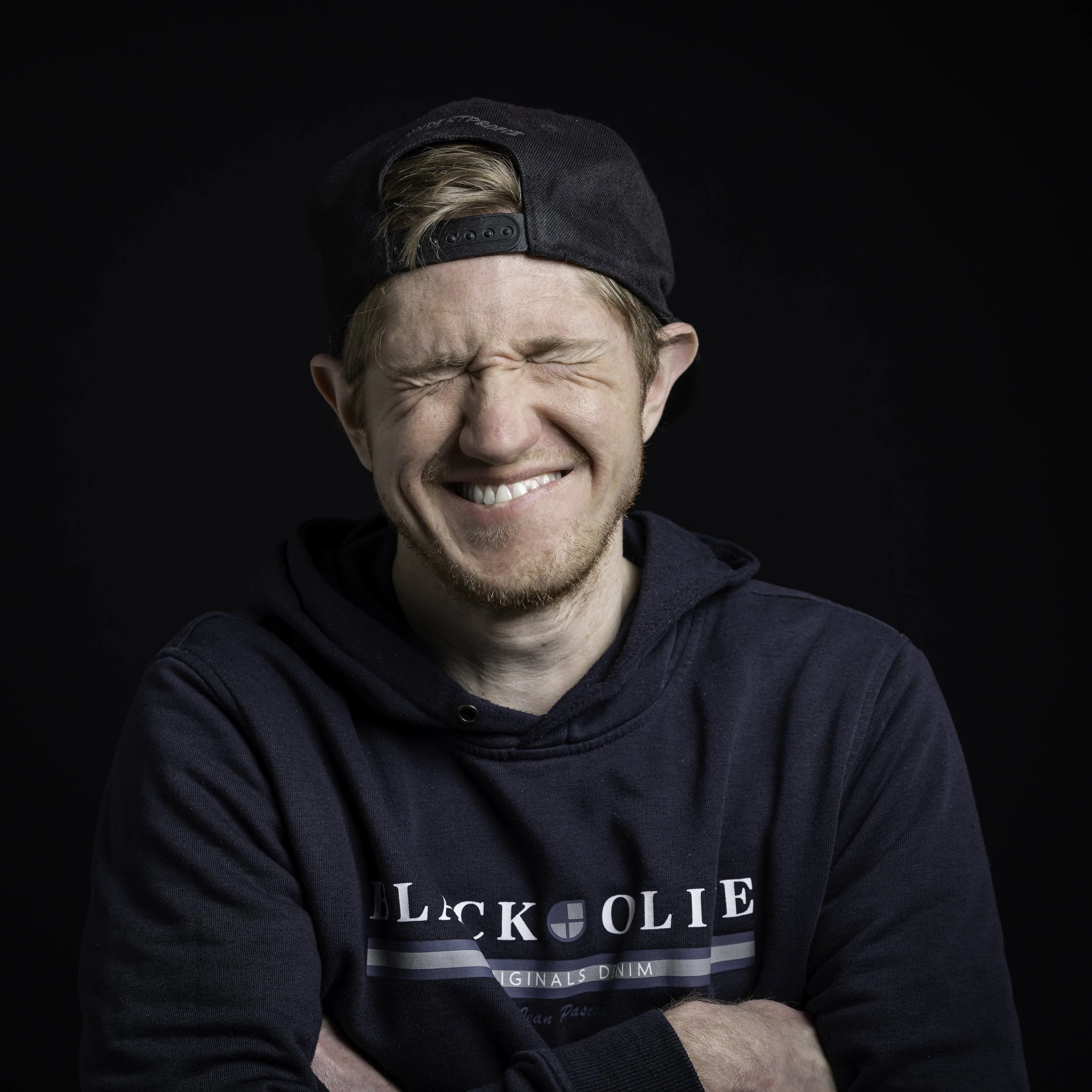 Mann trägt eine schwarze Baseballkappe, hat blondes Haar und ist mit verschränkten Armen abgebildet. Er trägt ein dunkelblaues Sweatshirt mit weißen Schriftzügen.