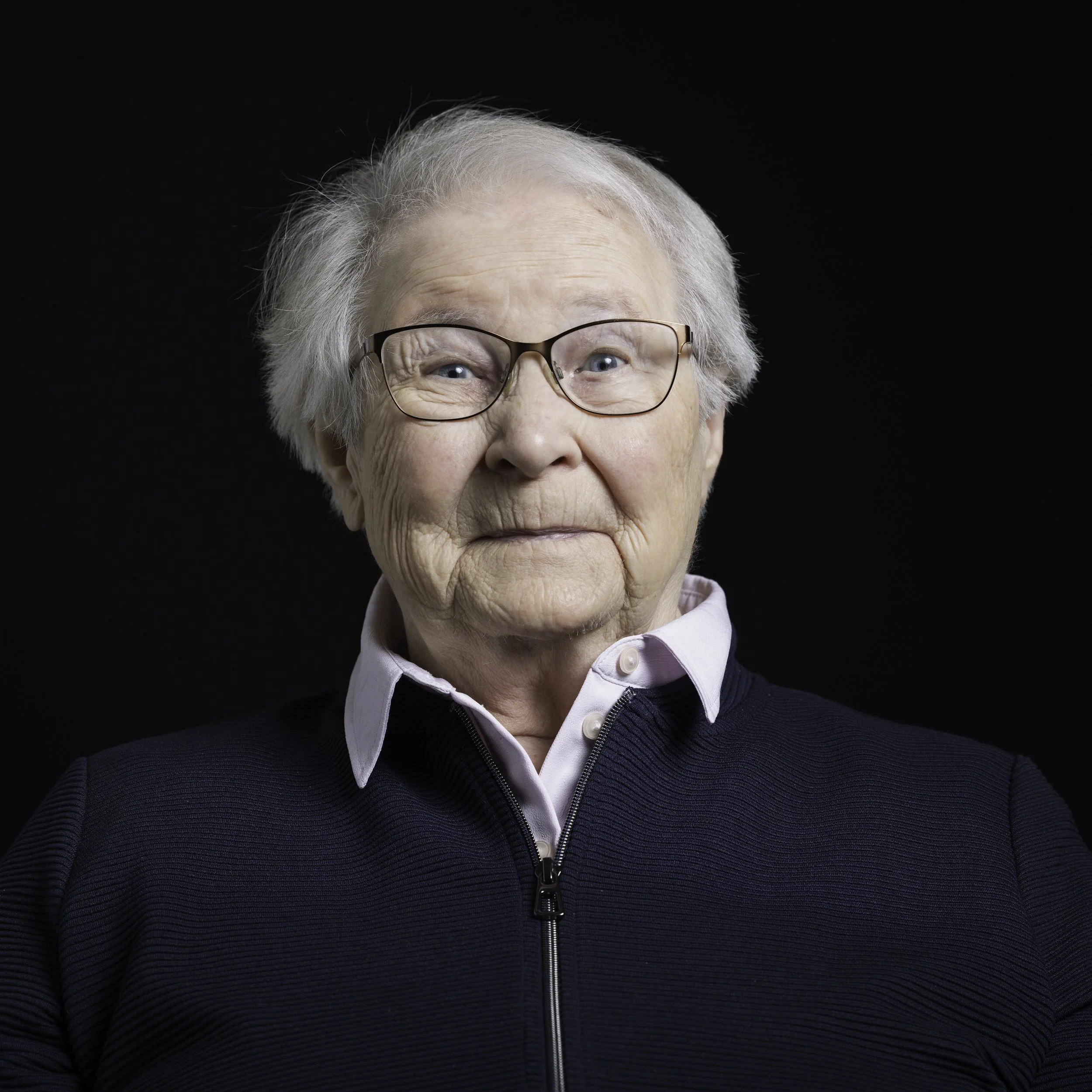 Portrait einer Dame mit weißem Haar, Brille und dunklem Pullover vor schwarzem Hintergrund.