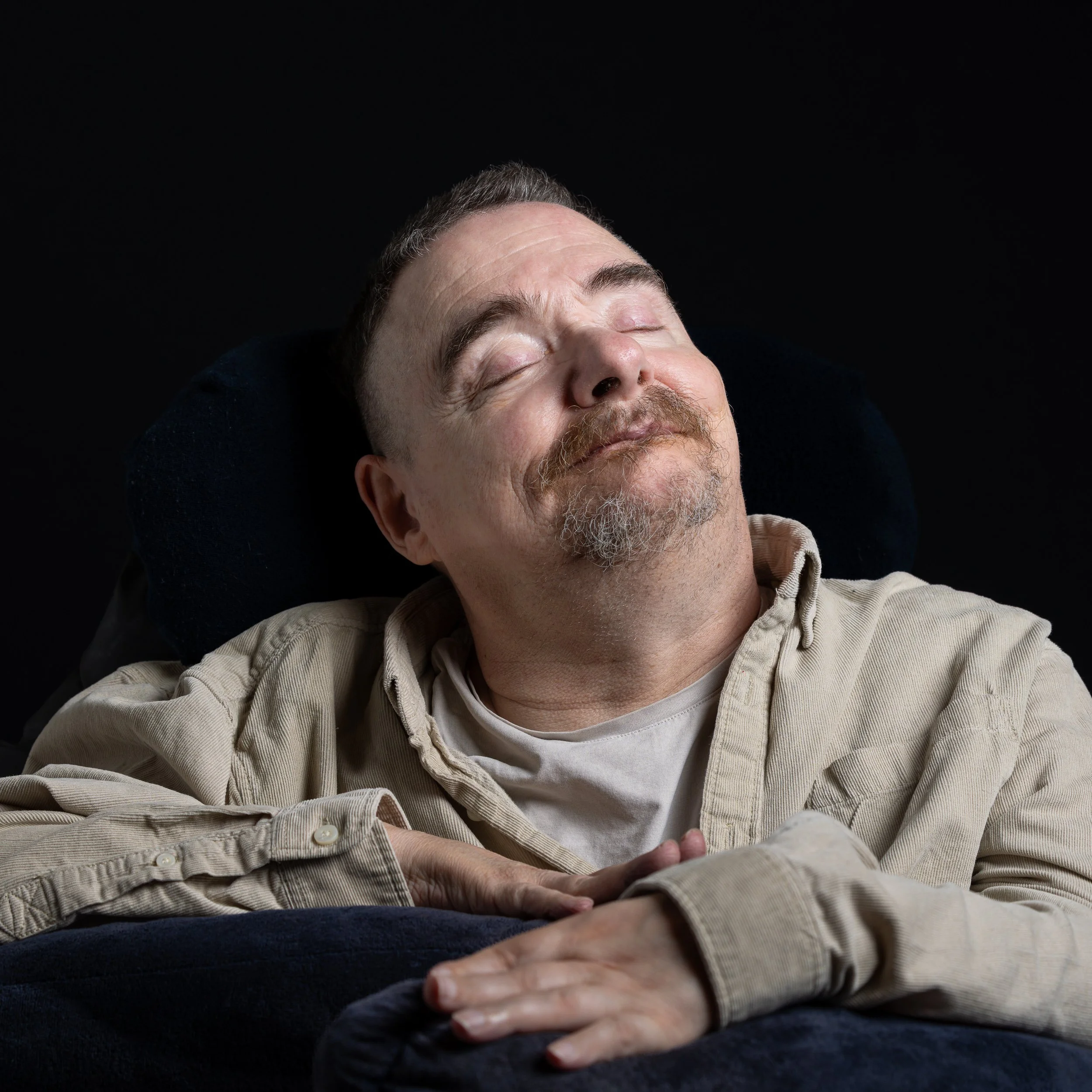 Ein Mann mit geschlossenen Augen liegt entspannt mit einem lächelnden Gesicht auf einem schwarzen Hintergrund, trägt ein beige-kariertes Hemd.