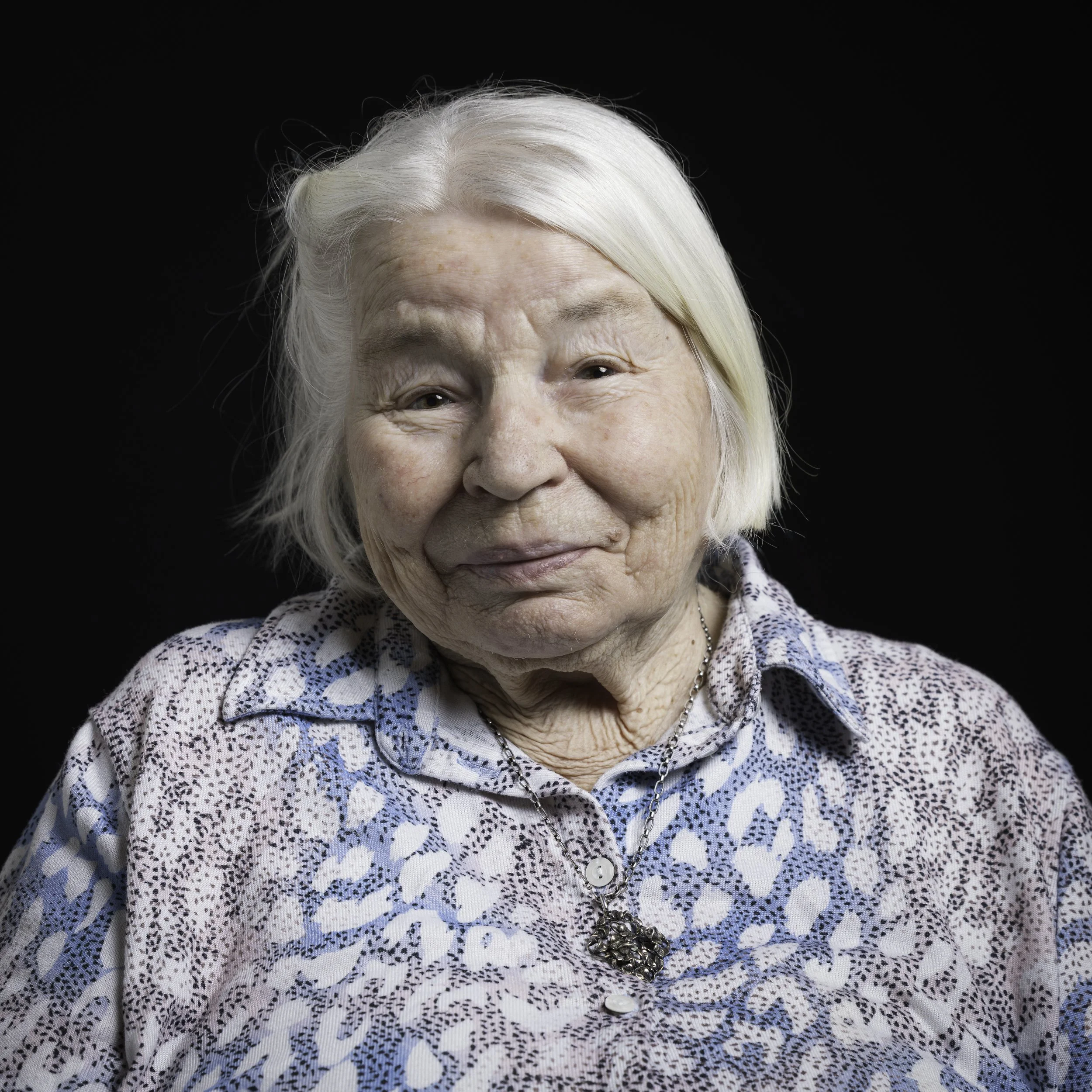 Nahaufnahme einer älteren Dame mit weißen Haaren und Bluse, gegen einen schwarzen Hintergrund.