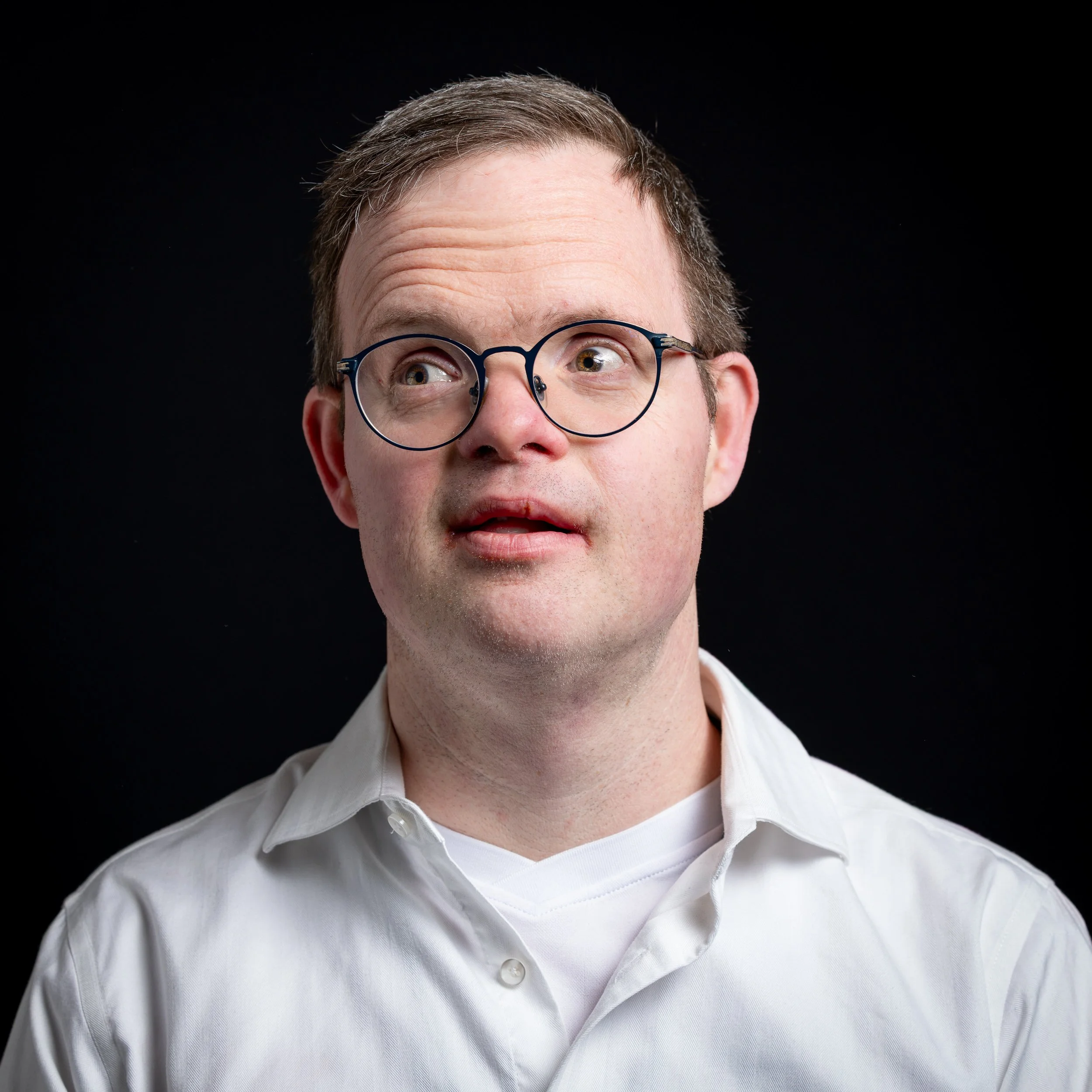 Nahaufnahme eines Mannes mit Brille, kurzer brauner Haarfrisur, trägt ein weißes Hemd, schaut nach oben, vor schwarzem Hintergrund.