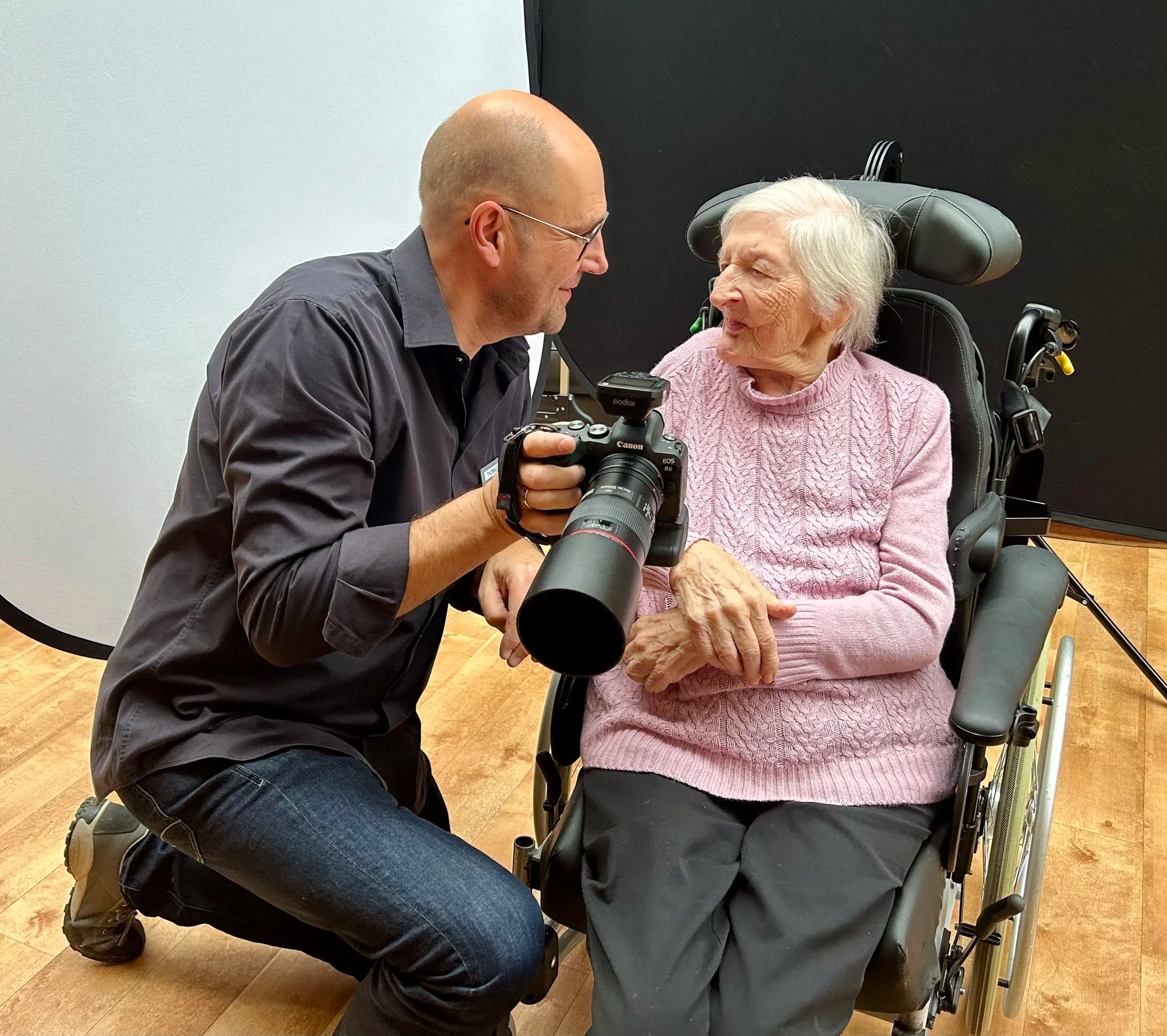 Fotograf Thomas Kamp kniet neben einer älteren Frau im Rollstuhl, beide lächeln sich an, während der Fotograf eine Kamera hält.
