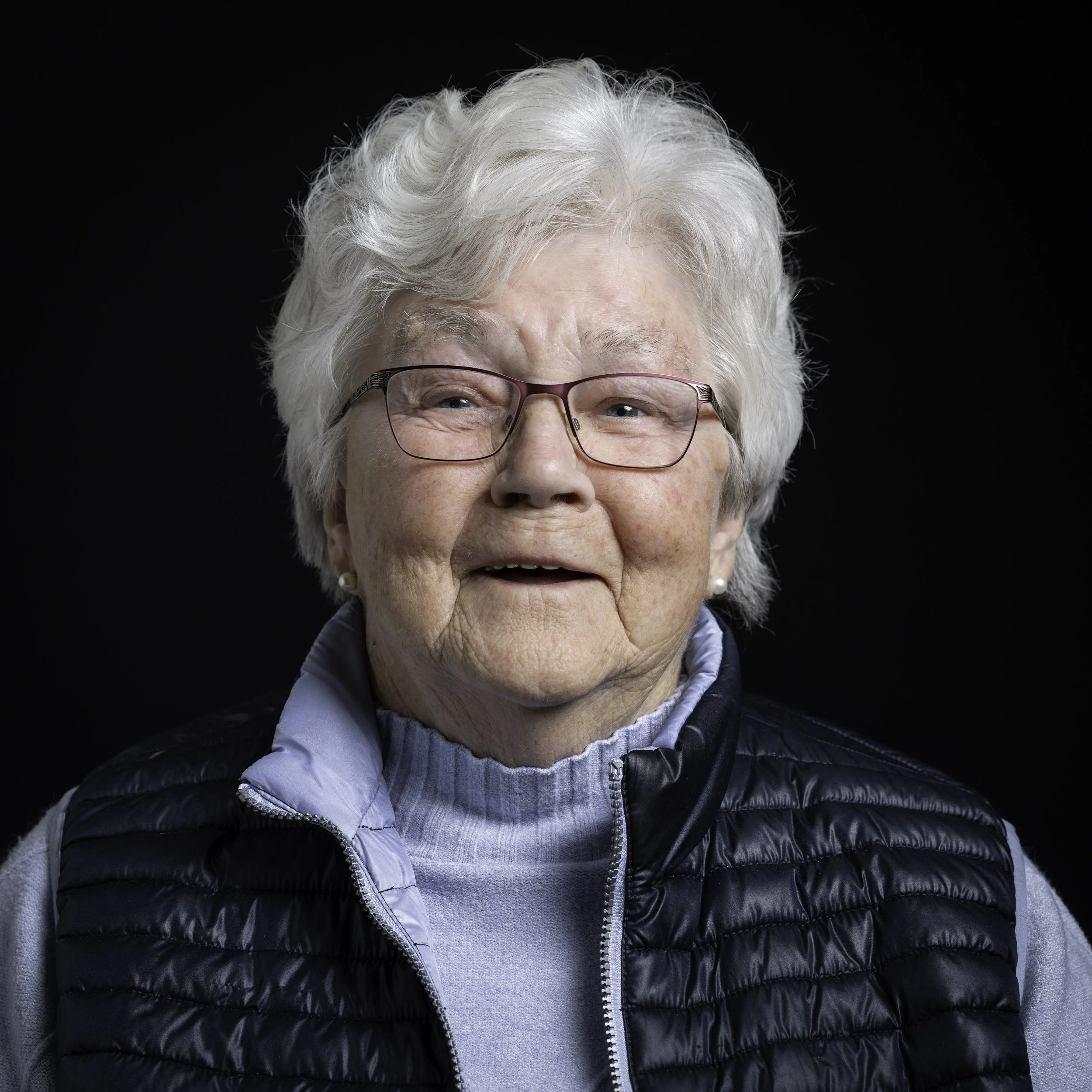 Nahaufnahme einer älteren Dame mit weißen Haaren, Brille und Lächeln, gegen einen schwarzen Hintergrund.
