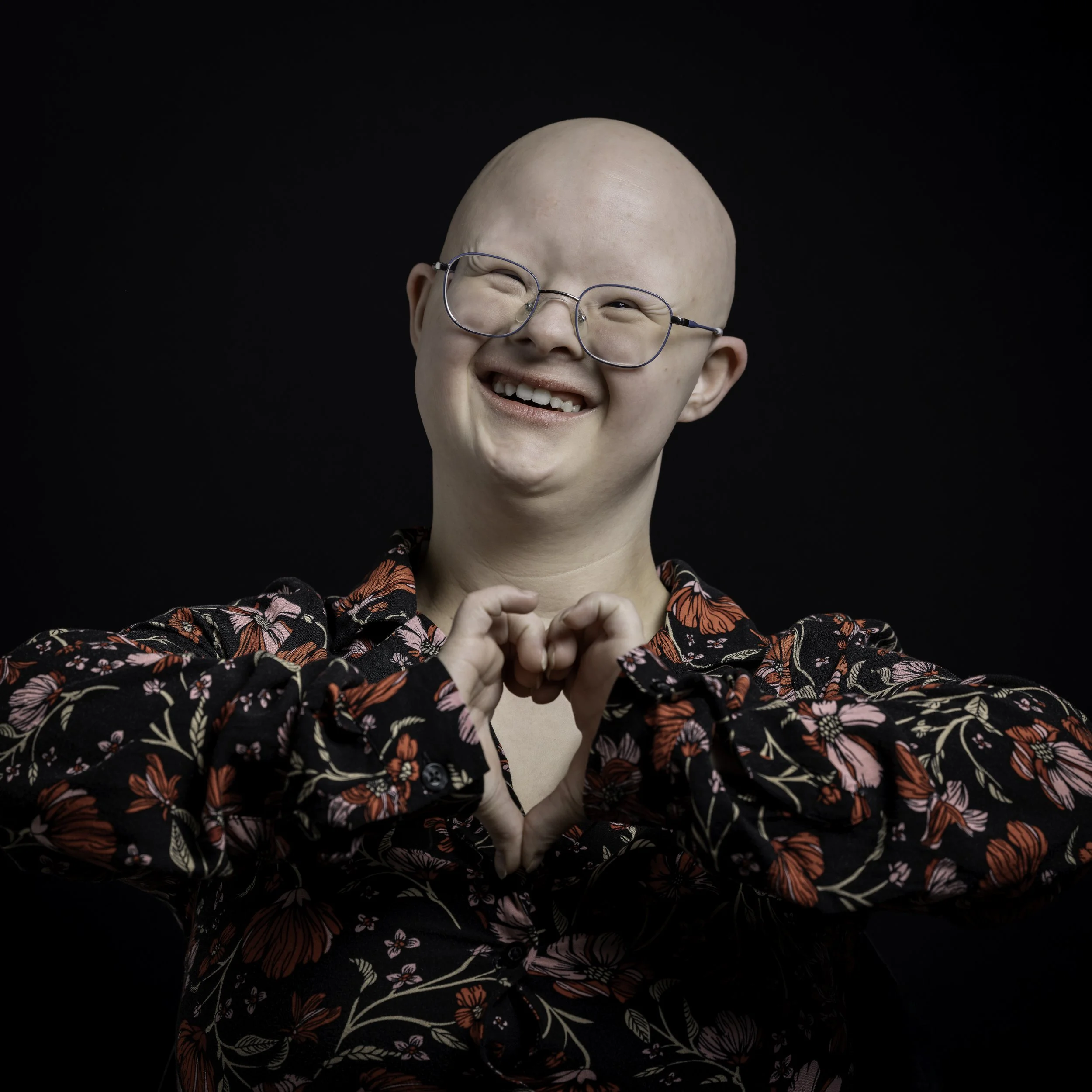 Ein lachender Mensch mit Brille, der eine Herzform mit den Händen bildet, vor dunklem Hintergrund.