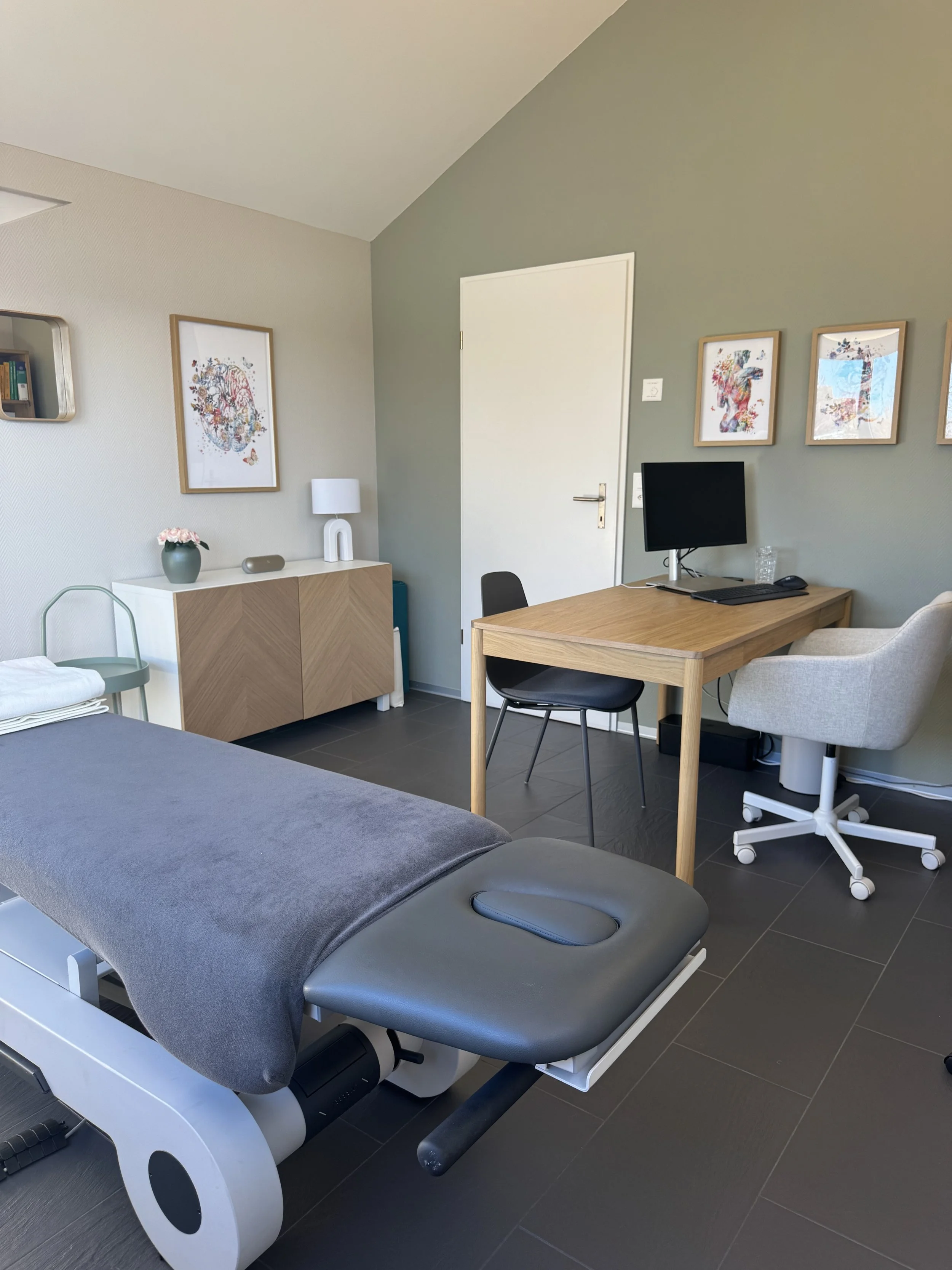 Behandlungsraum mit Untersuchungsliege, Schreibtisch mit Monitor, Stuhl und Krankenschwesterstuhl, Wanddekorationen, Sideboard, Lampe und Topfpflanze.
