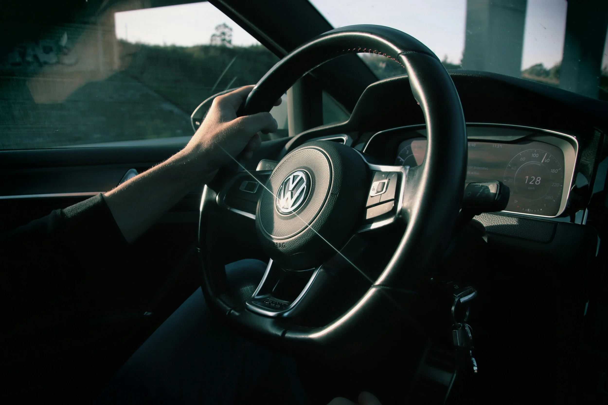 A person driving a Volkswagen vehicle, holding the steering wheel with their left hand, on a road during daytime.