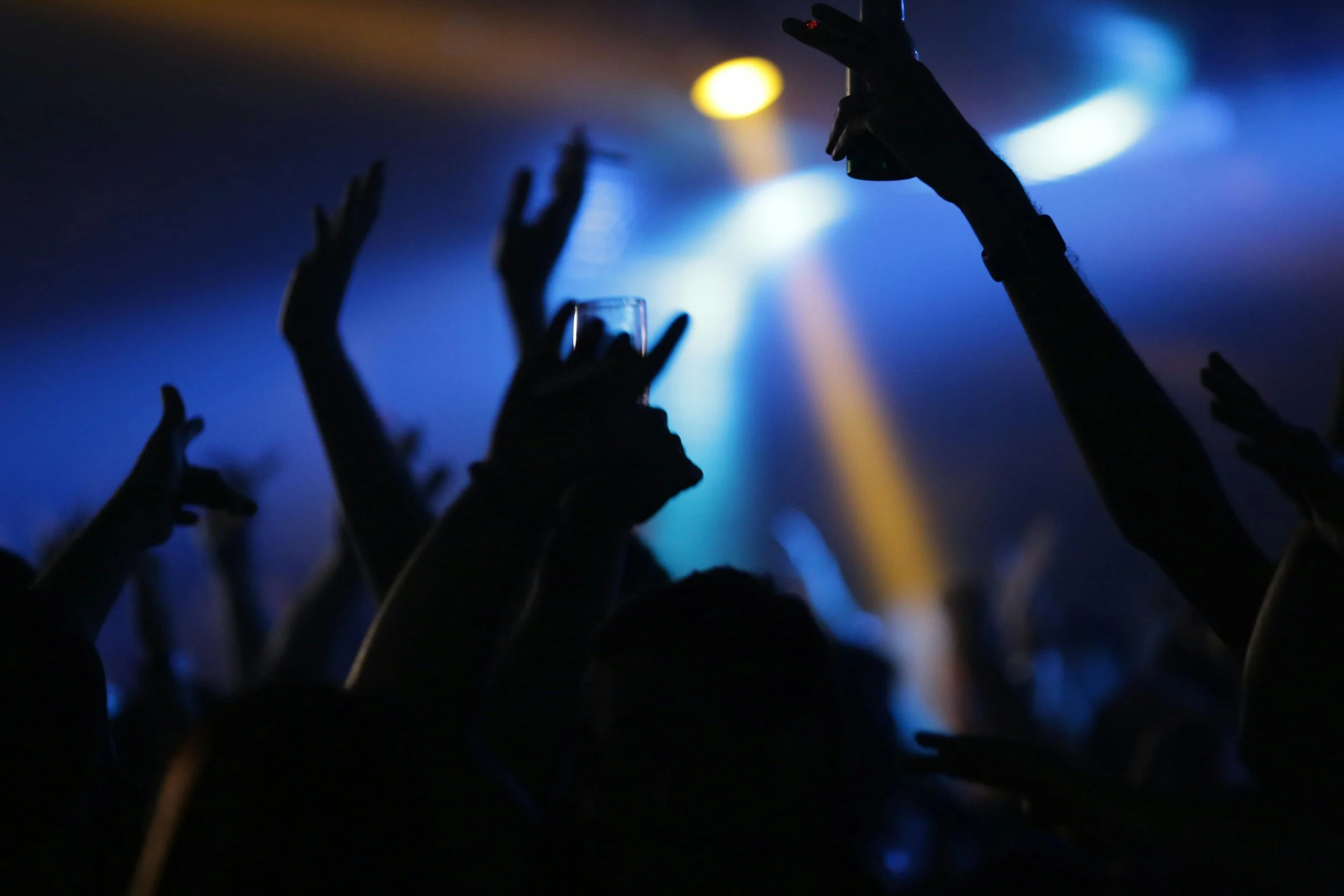 Silhouettes of people dancing with hands raised at a concert or nightclub, illuminated by blue and yellow stage lights.