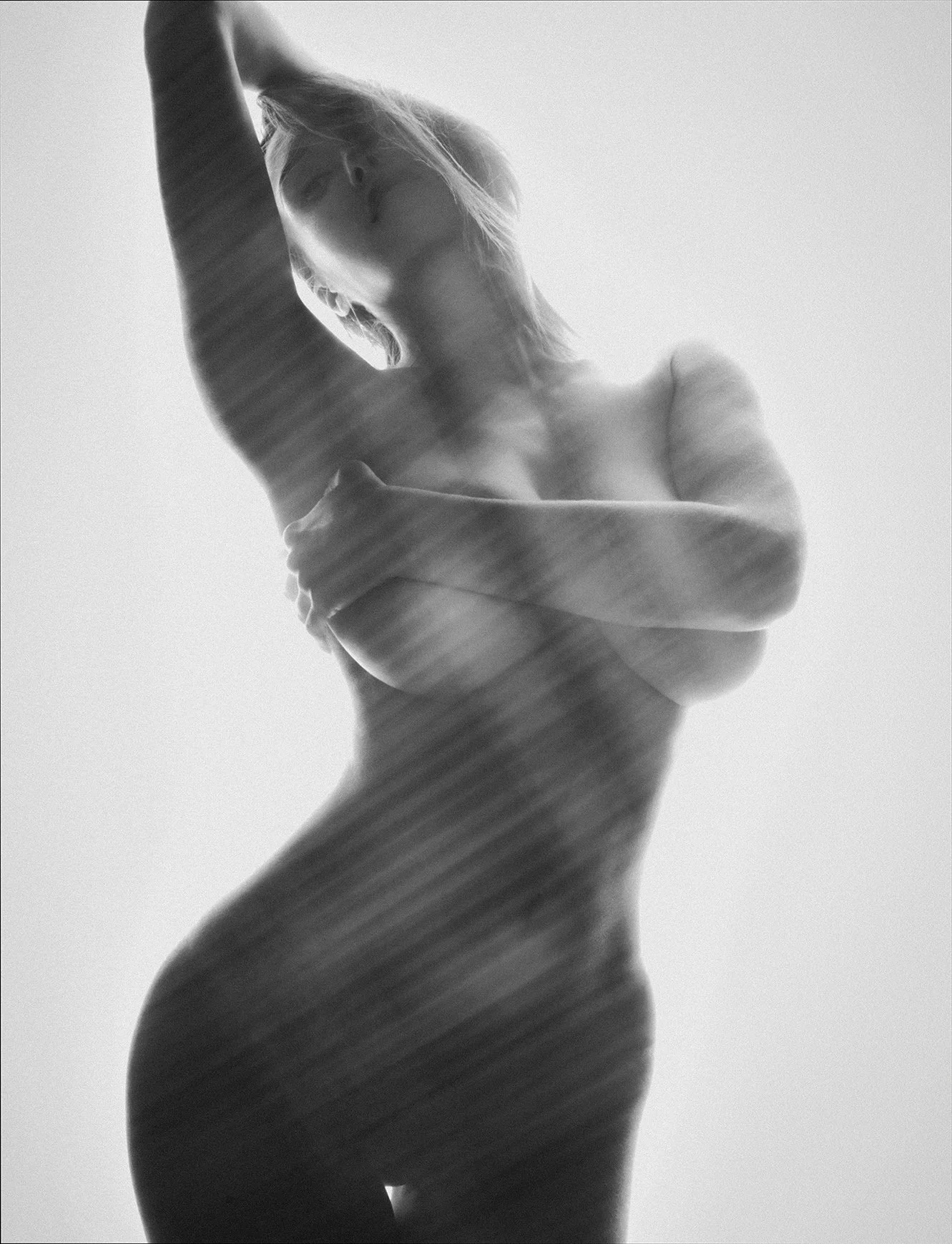 Black and white photograph of a woman posing topless, with light shining through, creating a silhouette effect. She has one arm raised above her head and the other covering her chest.