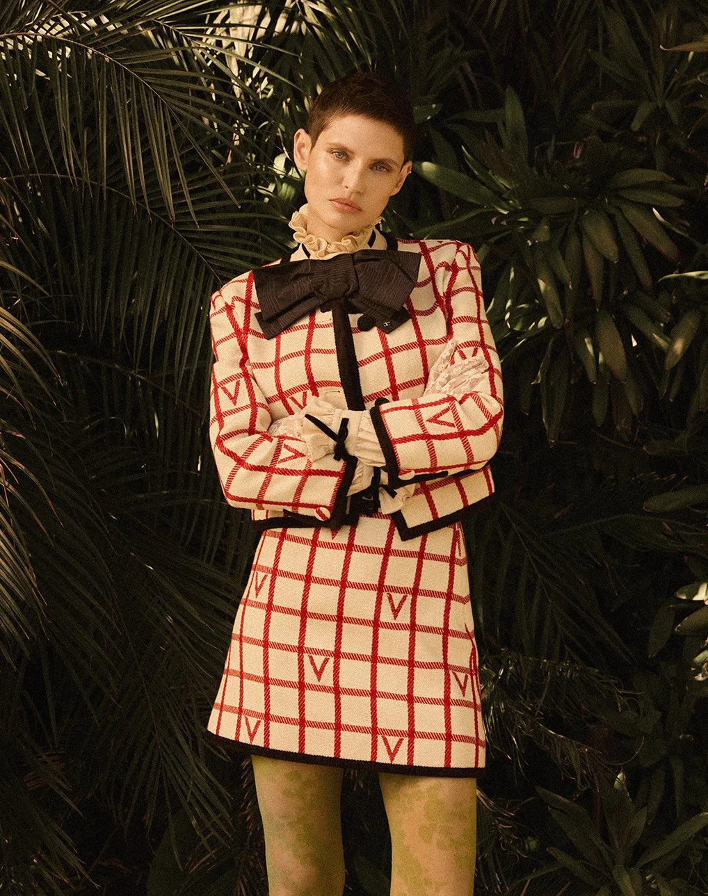 A woman with short dark hair and wearing a patterned outfit with a large bow, standing in front of lush green foliage.