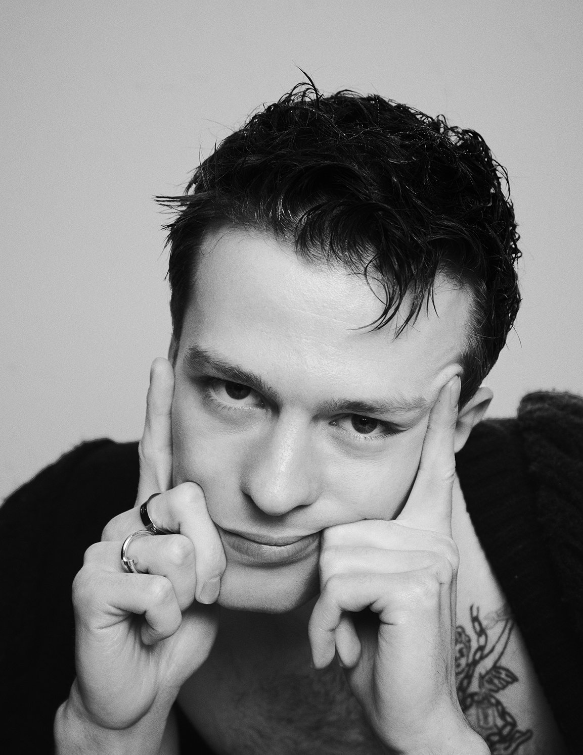 A black and white photo of a person with dark, wavy hair, holding their face with both hands, with a tattoo partially visible on their right arm.