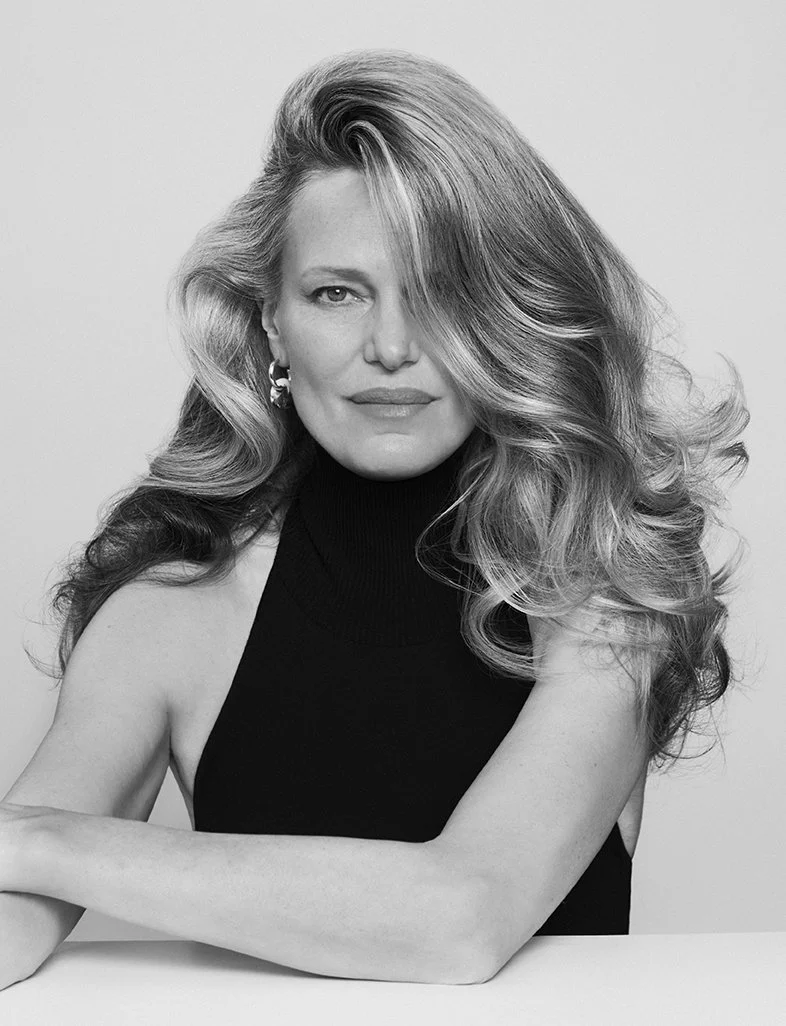 Black and white portrait of a woman with voluminous wavy hair, wearing a sleeveless black turtleneck top and earrings, sitting with her arms crossed, looking at the camera.