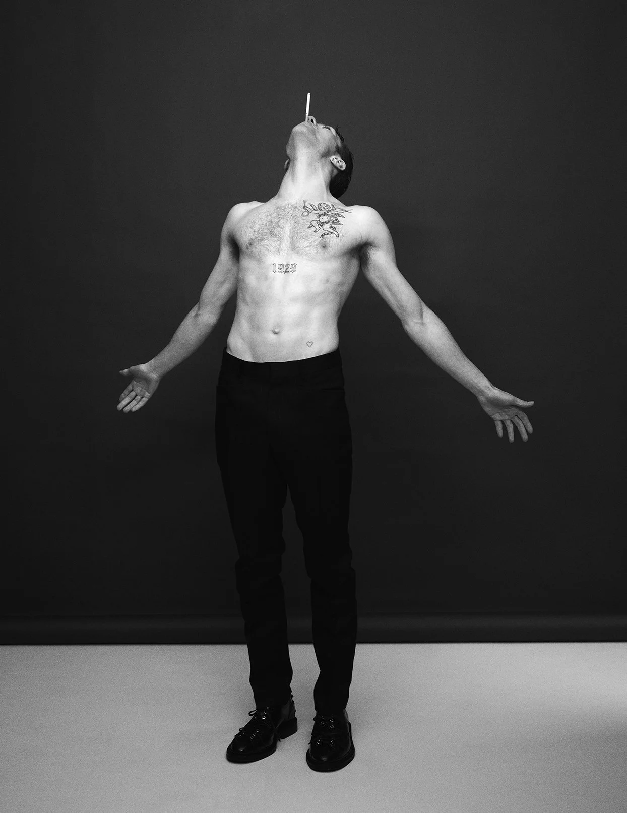 A shirtless man with tattoos stands with arms outstretched against a plain background, with a cigarette balanced on his nose.