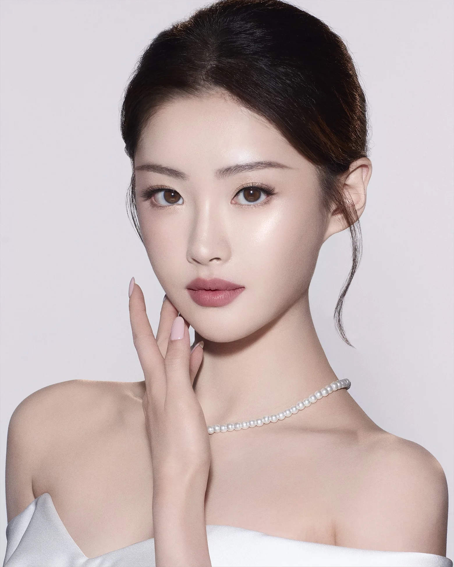 A close-up portrait of a young woman with fair skin, brown eyes, and dark brown hair styled with a slight wave. She is wearing a pearl necklace and a white off-the-shoulder top, with her right hand gently touching her face.