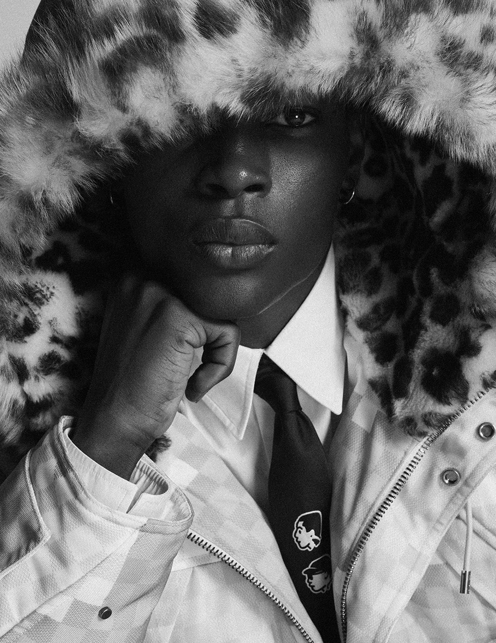 A black woman wearing a patterned furry hooded coat and a white shirt with a tie, resting her chin on her hand, looking into the camera.