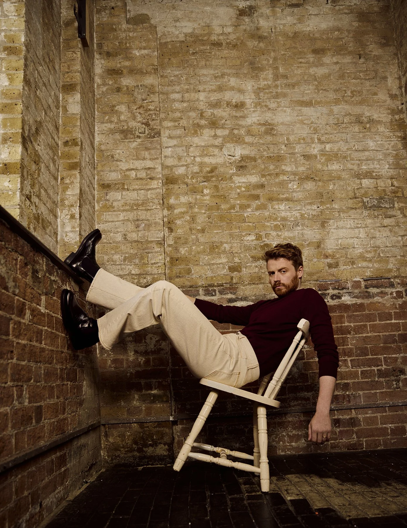 A man sitting on a small white chair with his legs raised and resting against a brick wall in a room with a brick wall backdrop.