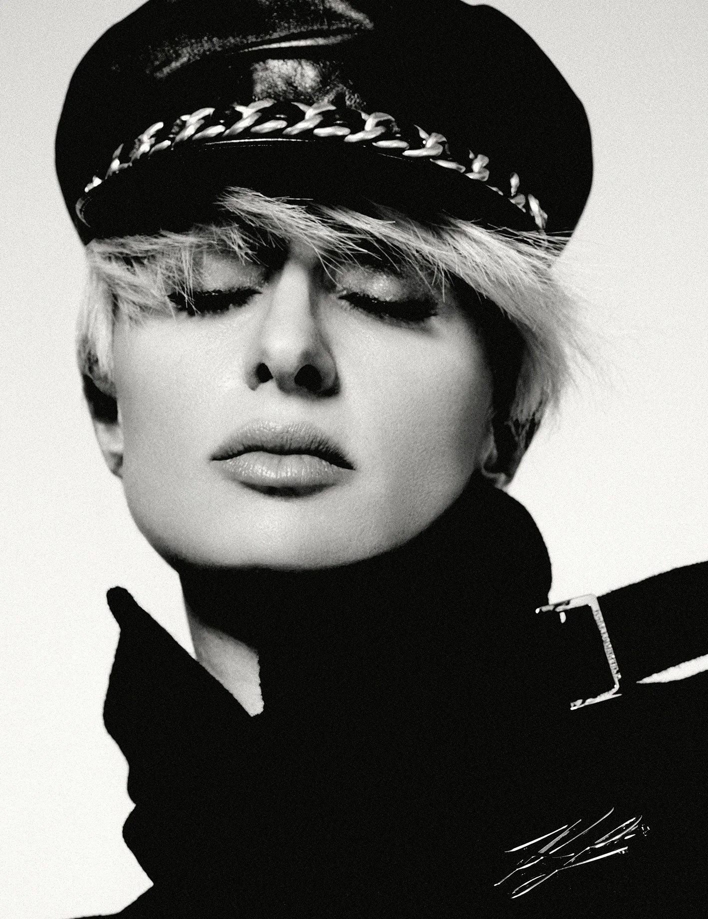 A black-and-white portrait of a woman with short blonde hair, wearing a black hat with a chain detail and a black high-collared outfit with a zipper.