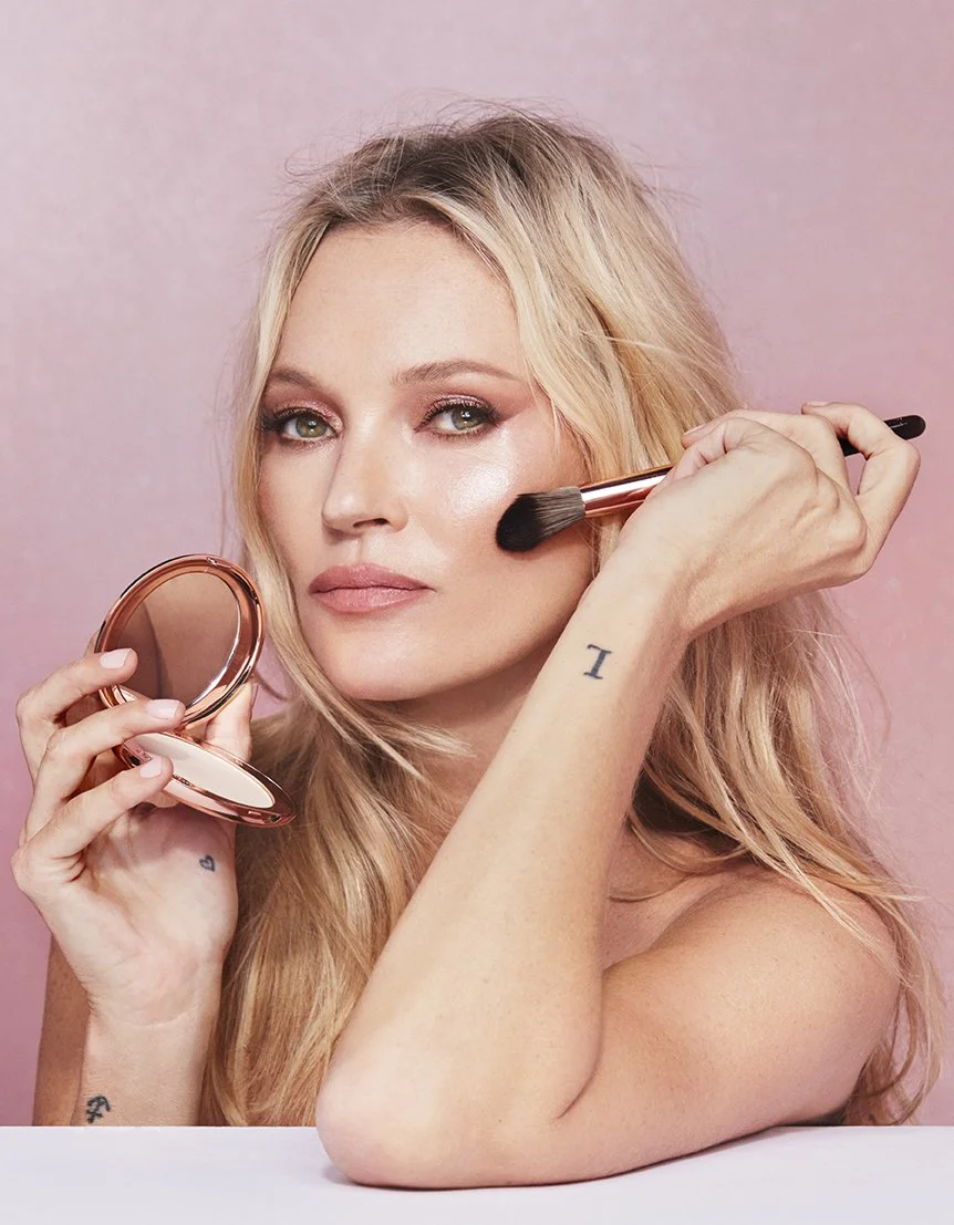 A woman with blonde hair applying blush or highlighter with a makeup brush while holding a compact mirror against a pink background.