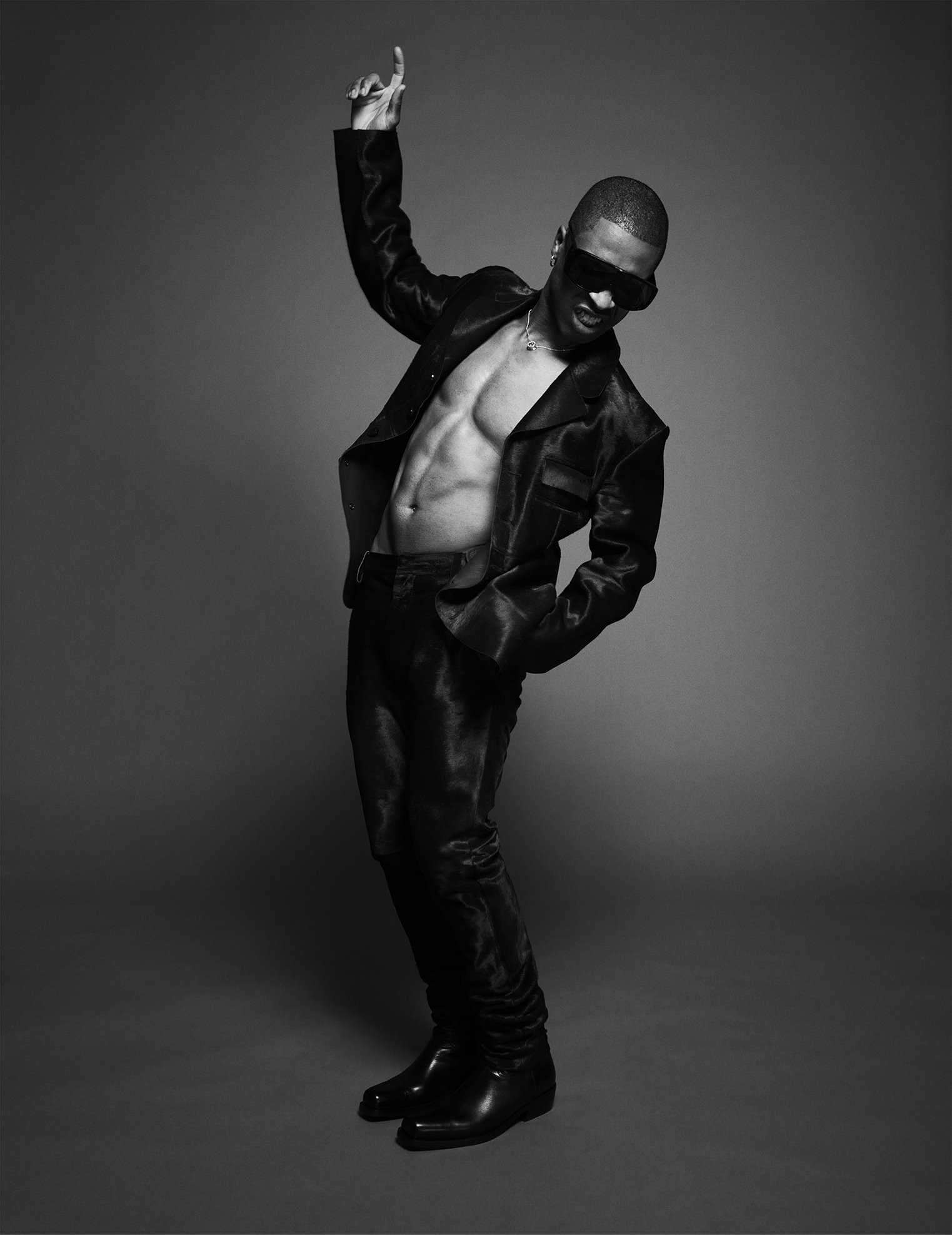 Black and white photo of a man wearing sunglasses, an open shiny black jacket, shiny black pants, and boots, posing with one arm raised and the head turned downward.