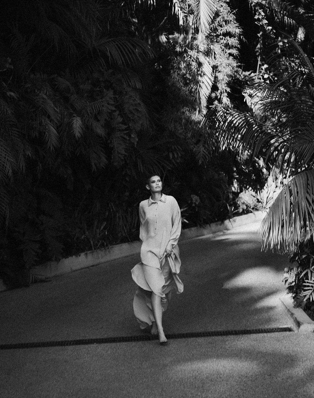 A woman in a long dress walking on a paved path surrounded by lush tropical plants.